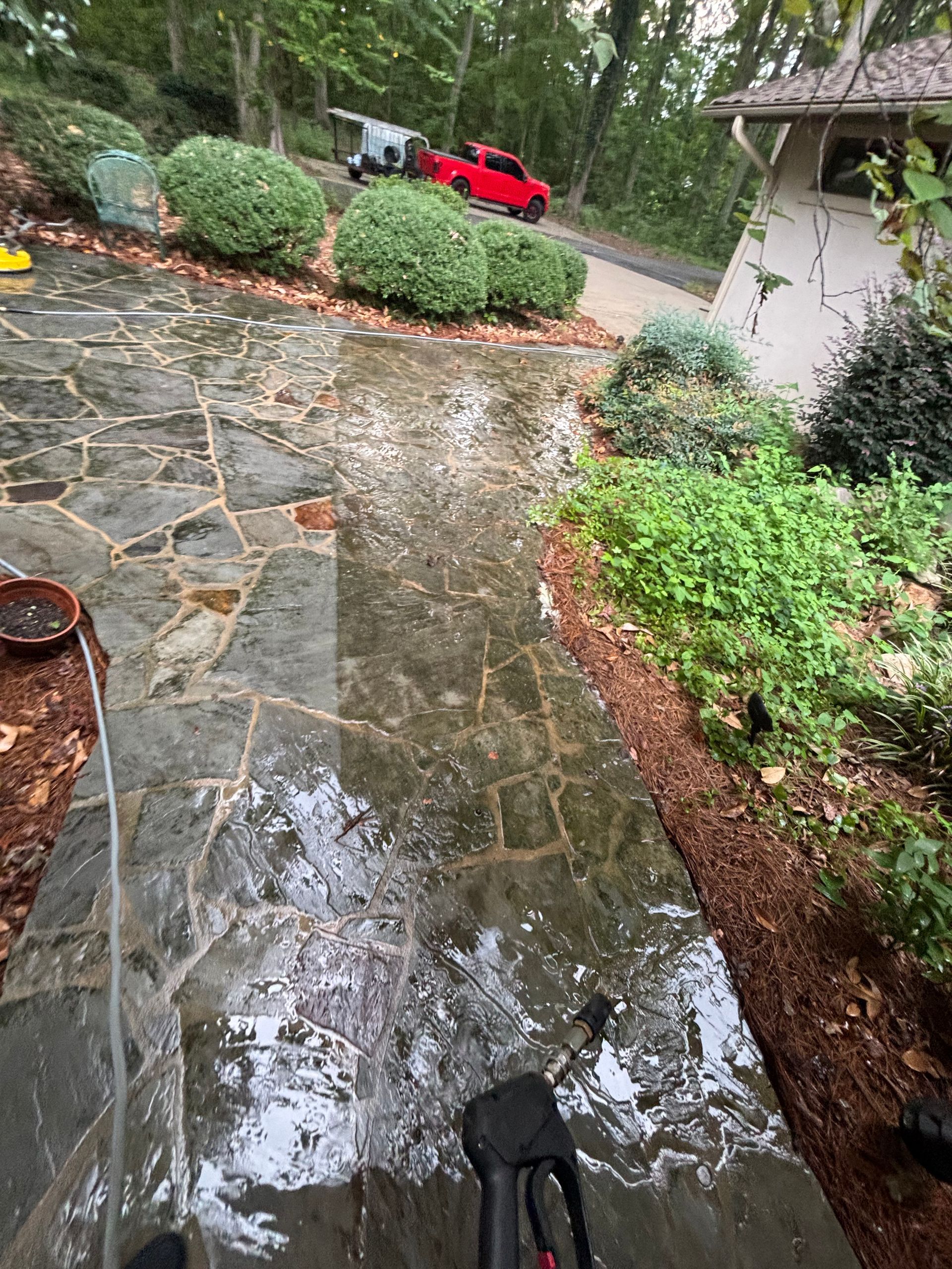Wet stone patio being power washed, showing a clean vs. dirty surface, with landscaping in the background.