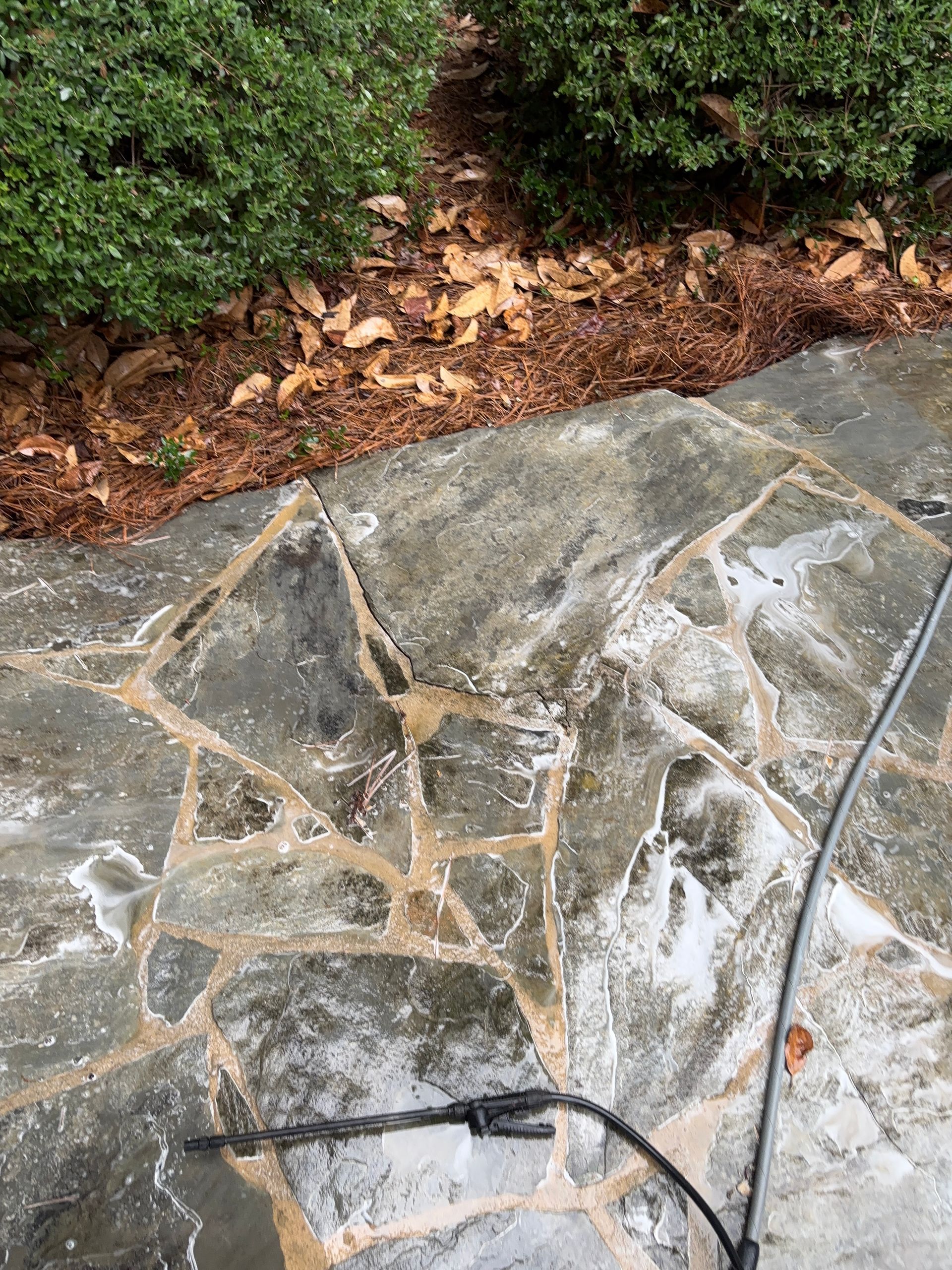 Stone patio being cleaned with a pressure washer near a bed of mulch and green shrubs.