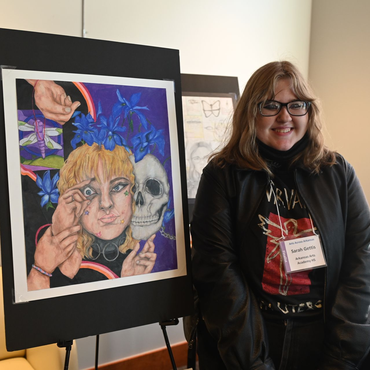 Woman with glasses smiles next to her artwork, a surreal painting of a face, skull, and hands.