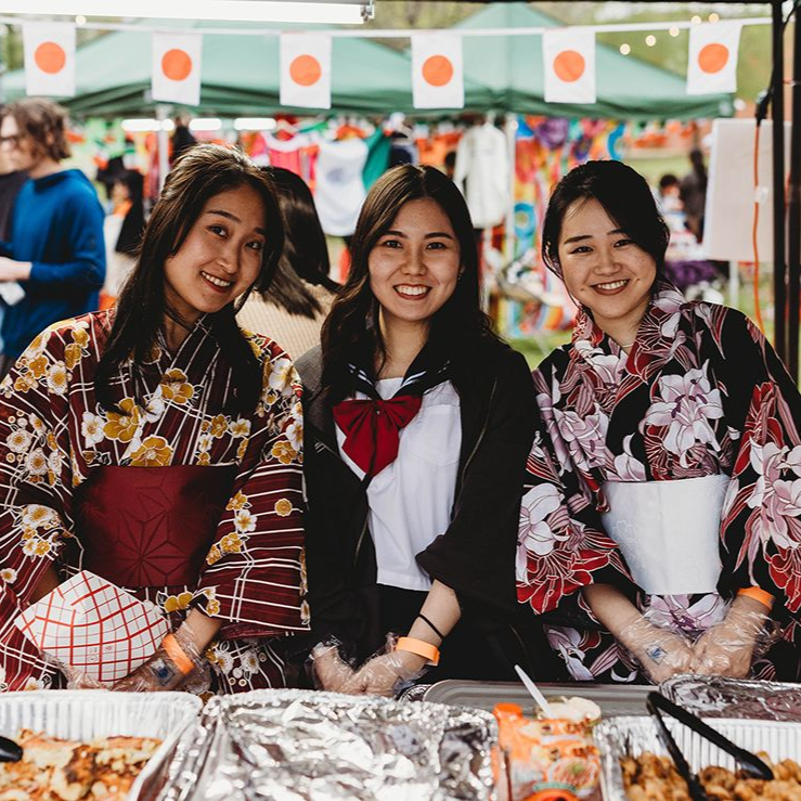 Three women at a Japanese food stall. Two wear floral kimonos, one a school uniform. Outdoor event, flags.