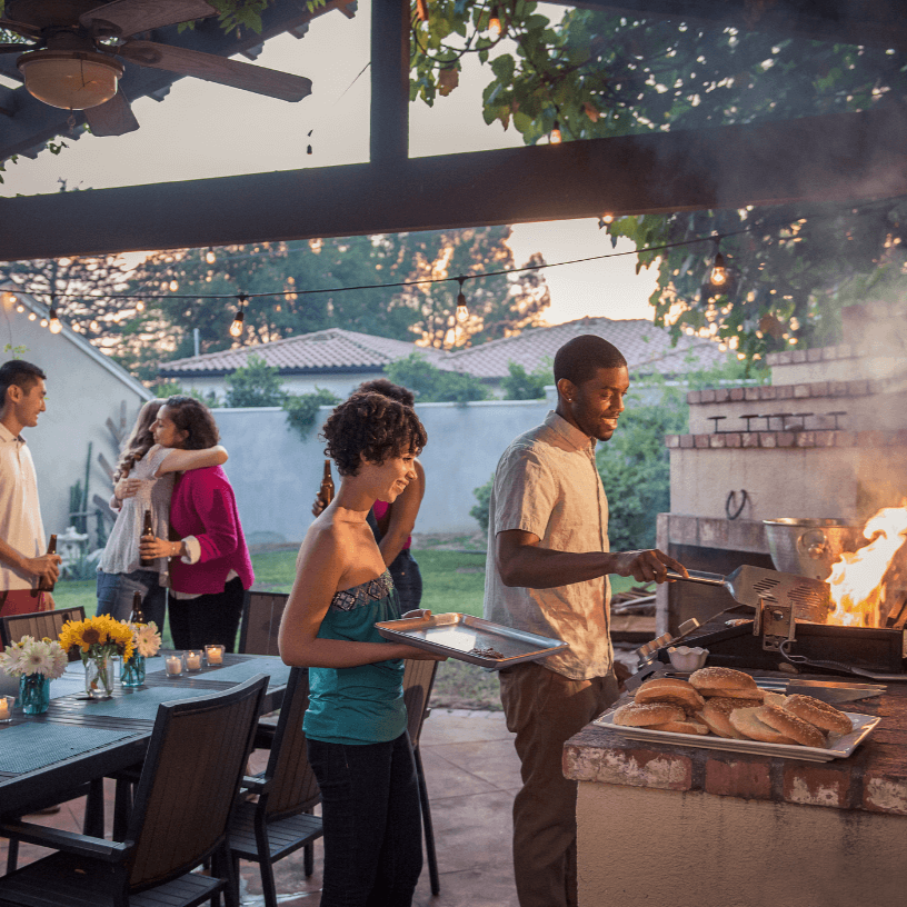 Group of people at a backyard barbecue with a grill, food, and string lights at dusk.