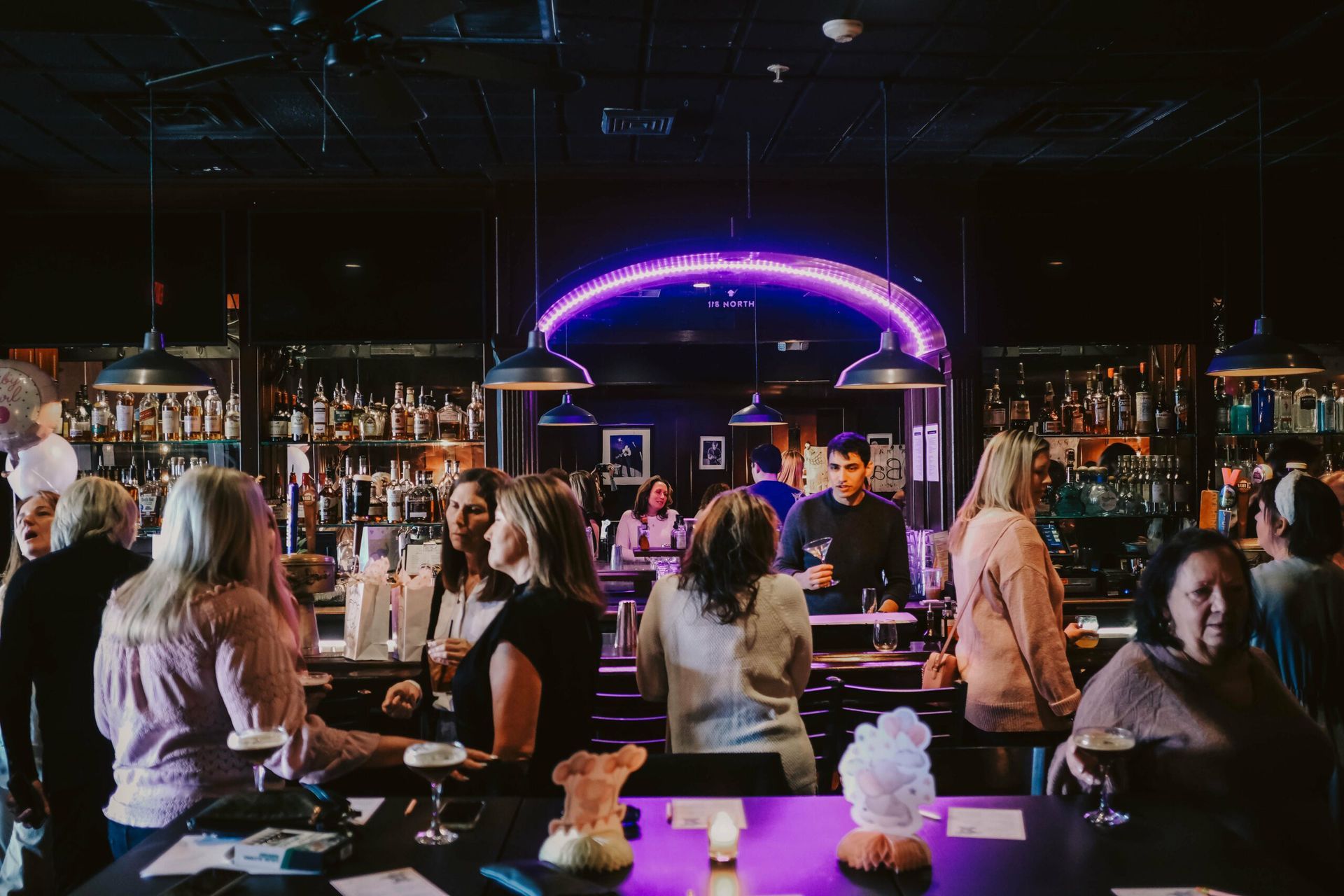 Bar scene with people around a dark bar, purple neon arch, bartender making drinks.
