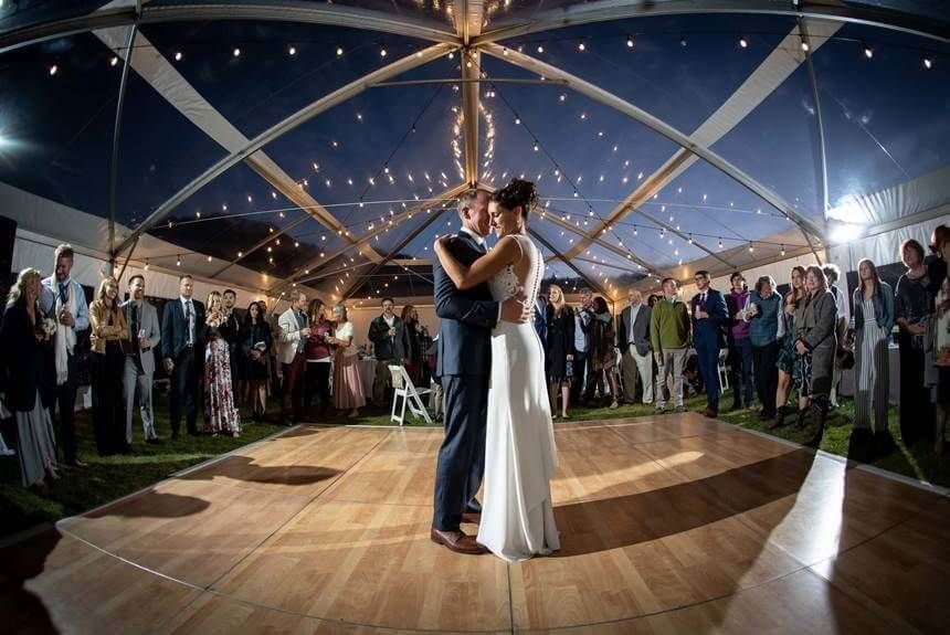 Couple dances under a clear tent, surrounded by wedding guests. Strings of lights are overhead.