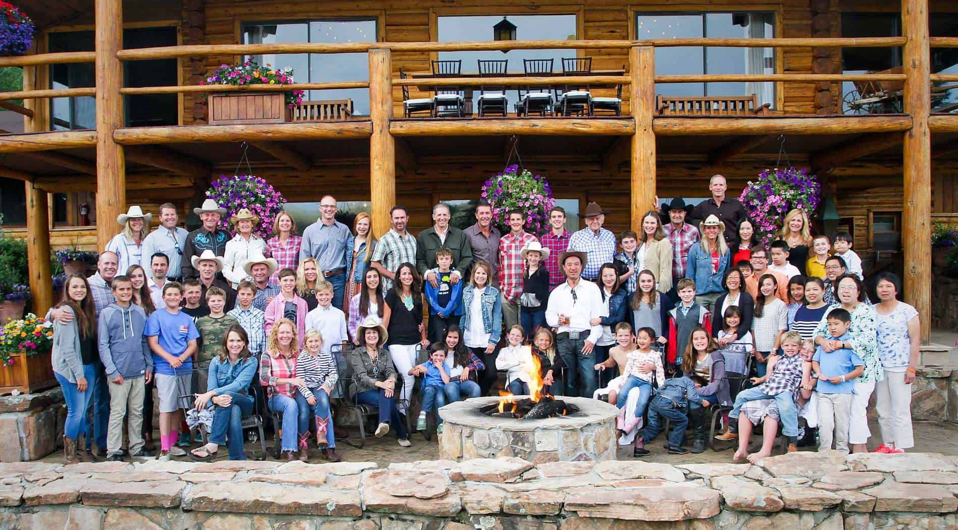 Large group of people posing in front of a log cabin with a stone fire pit; many are smiling.