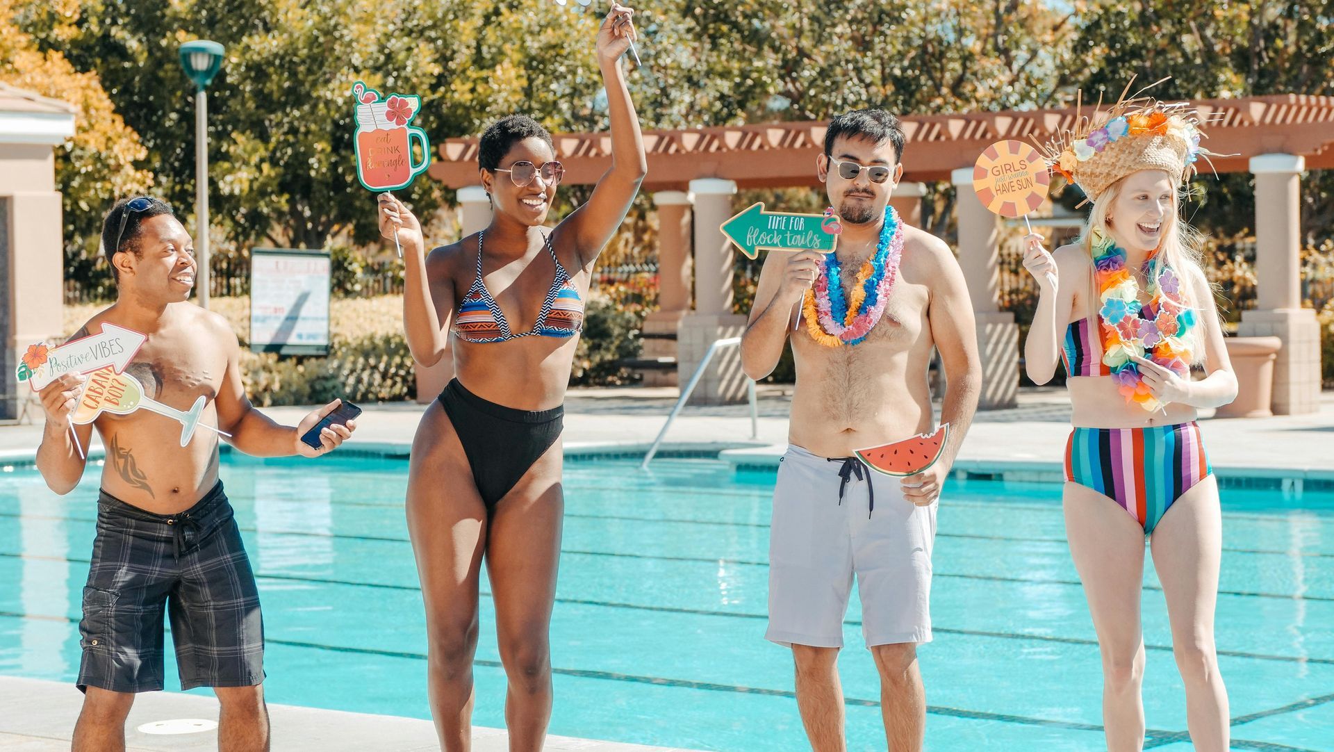 Four people in swimwear by a pool; holding tropical props, smiling, celebrating. Sunny day.