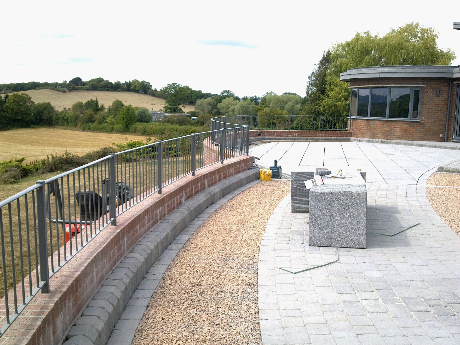 Curved brick patio with metal railing overlooking a field. Building with windows on the right, gravel border.