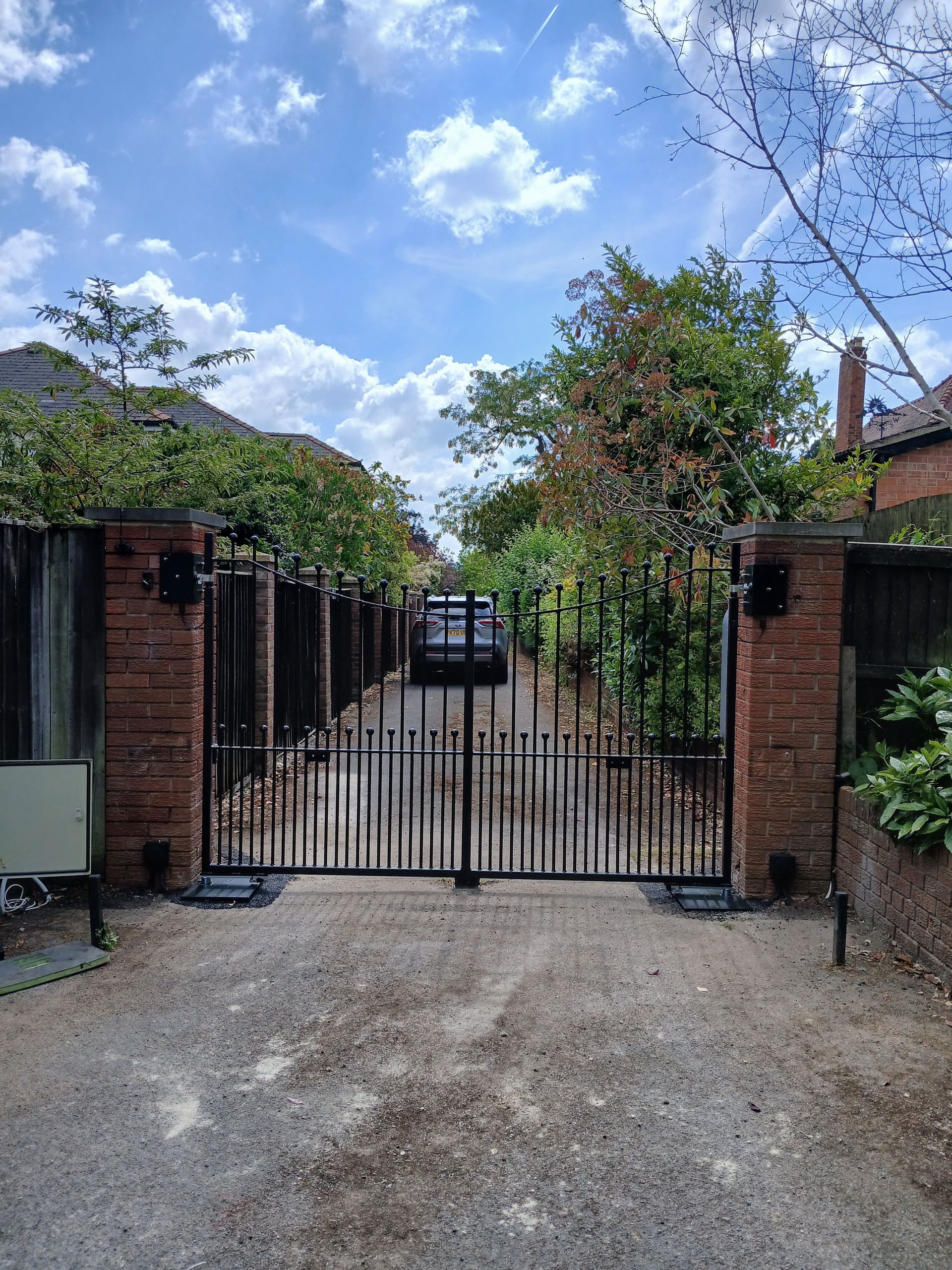 Black wrought iron gates open to a gravel driveway. Red brick pillars and a car are visible in the distance.