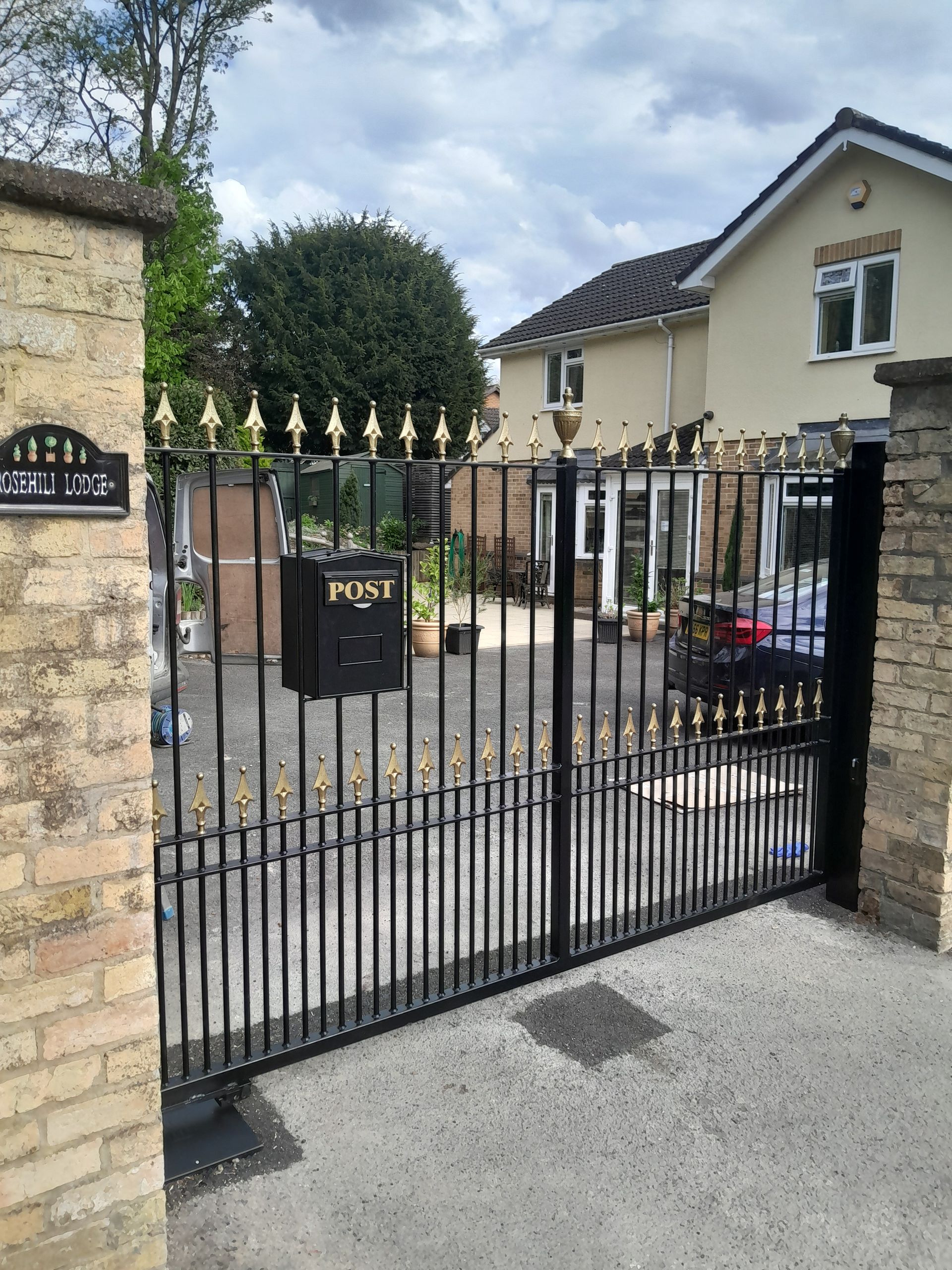 Black wrought iron gates with gold accents, mailbox, leading to a house, overcast day.