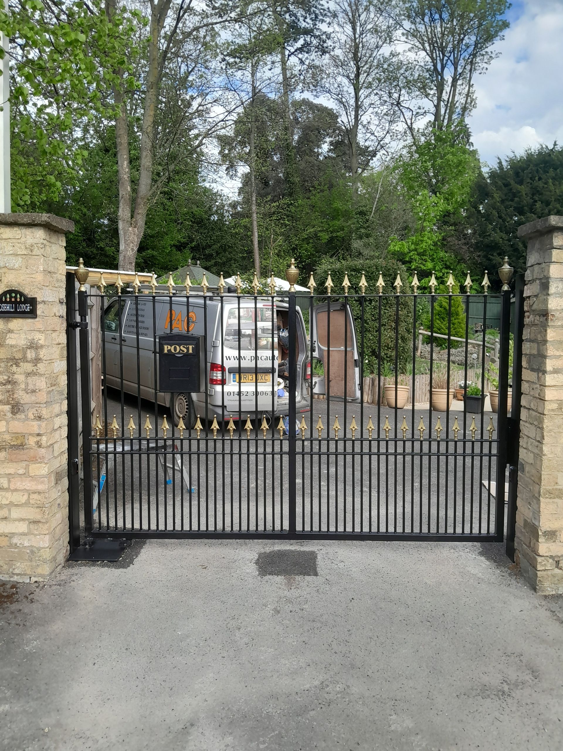 Black wrought iron gate with gold accents, brick pillars, driveway, van parked behind the gate, and trees in the background.