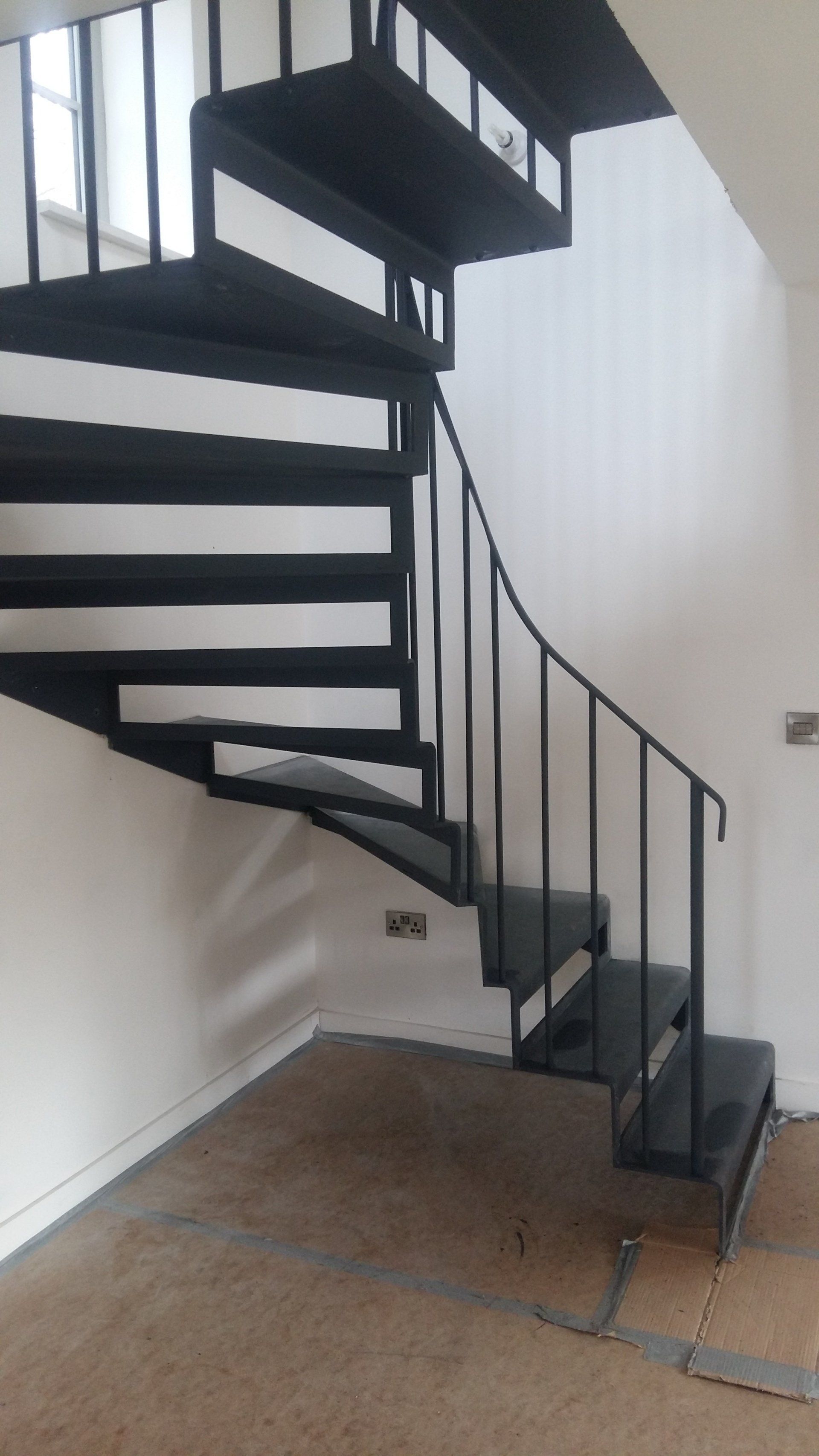 Black spiral staircase with metal railing and steps in a room with white walls and a brown floor.