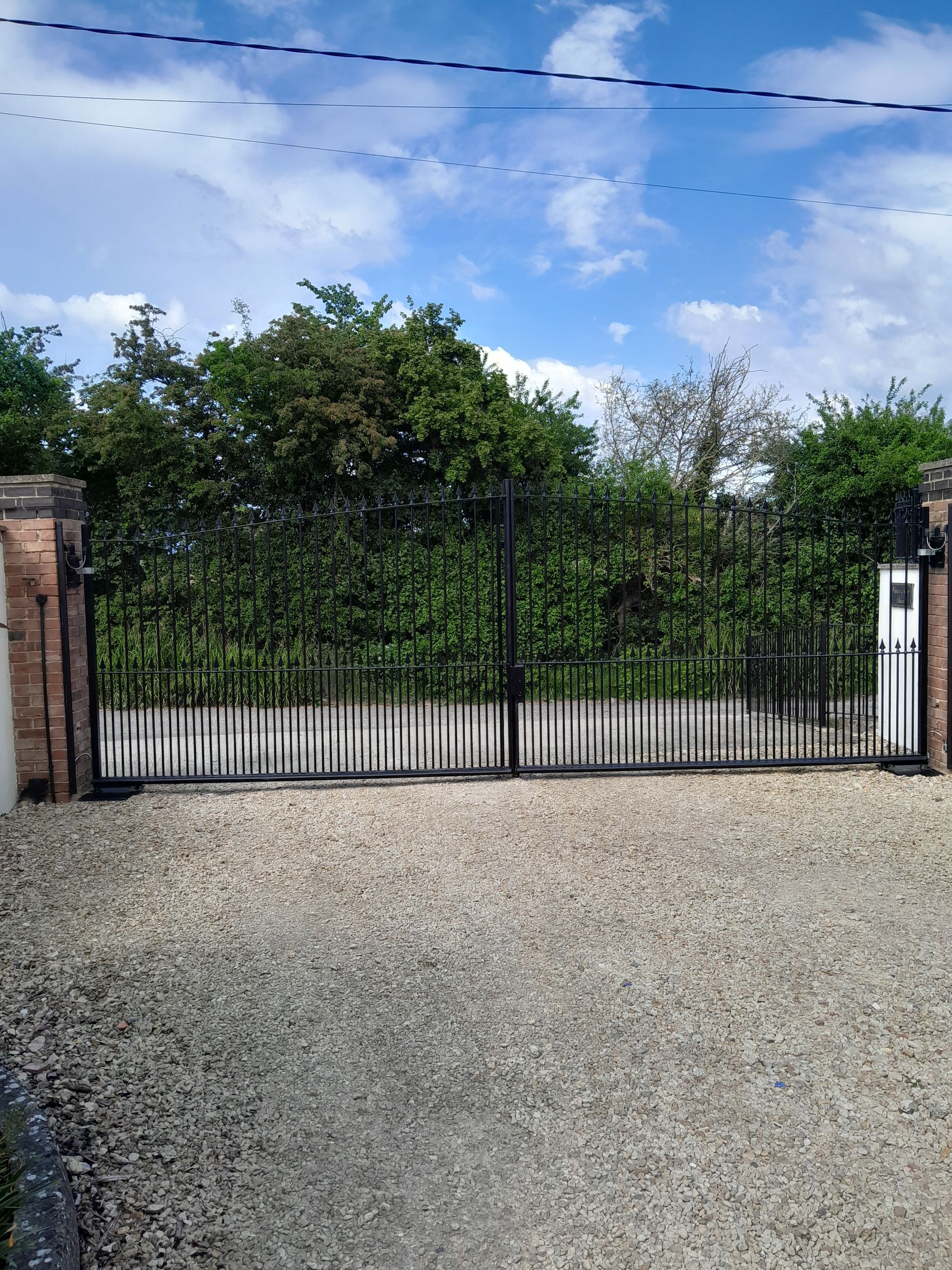 Black wrought iron gates open onto a gravel driveway; brick pillars, green foliage, blue sky.