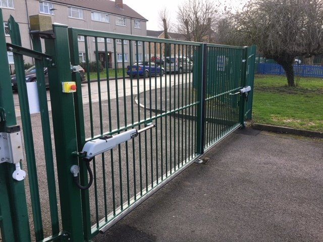 Green metal gate with vertical bars, driveway, and automatic opener.