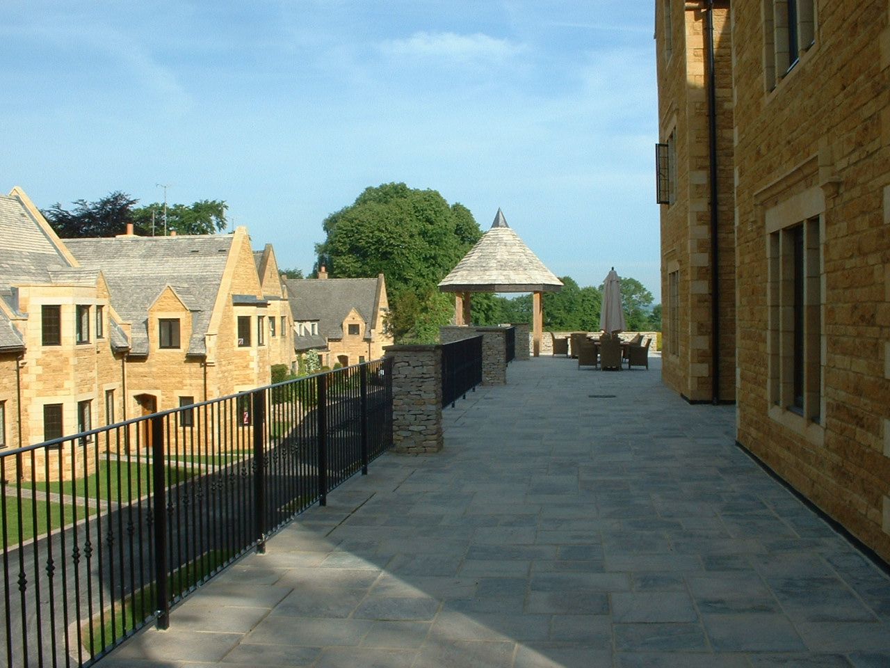 Exterior patio with stone paving, black railings, and buildings with stone facades, under a blue sky.
