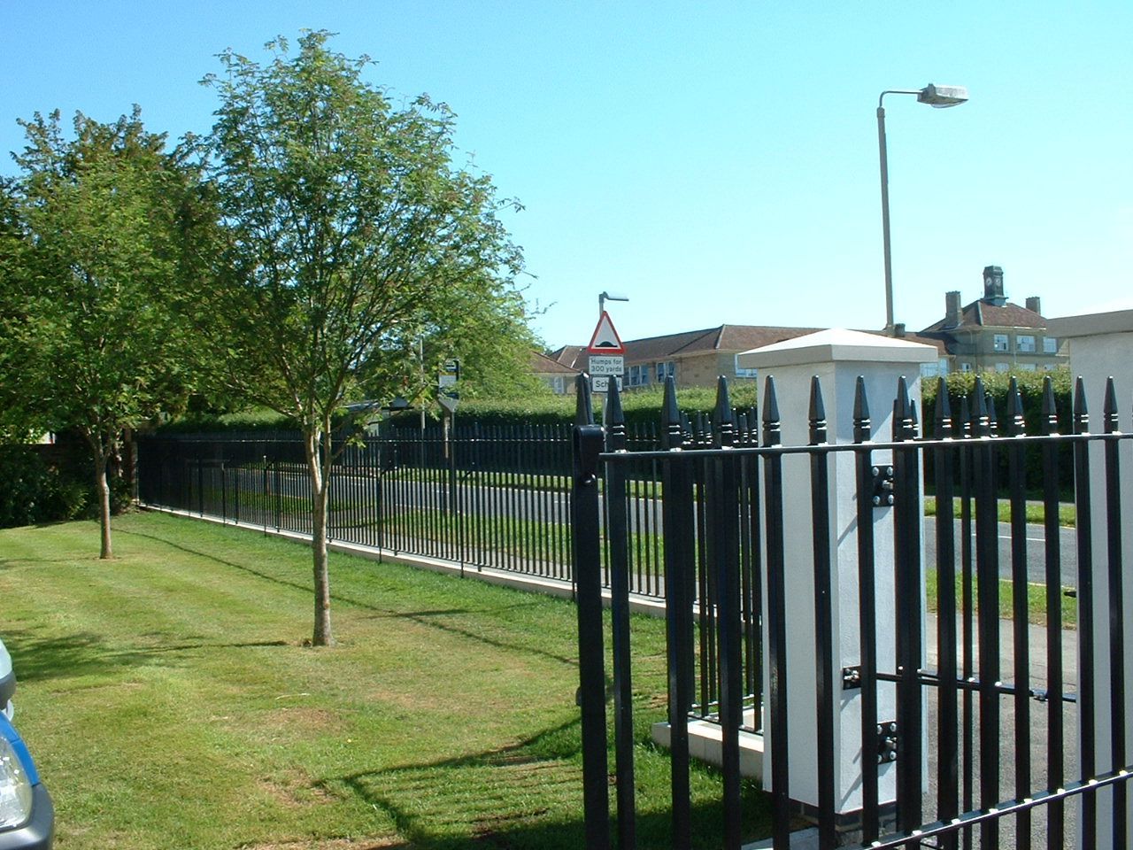 Black metal fence along a grassy lawn, trees, and buildings under a clear blue sky.