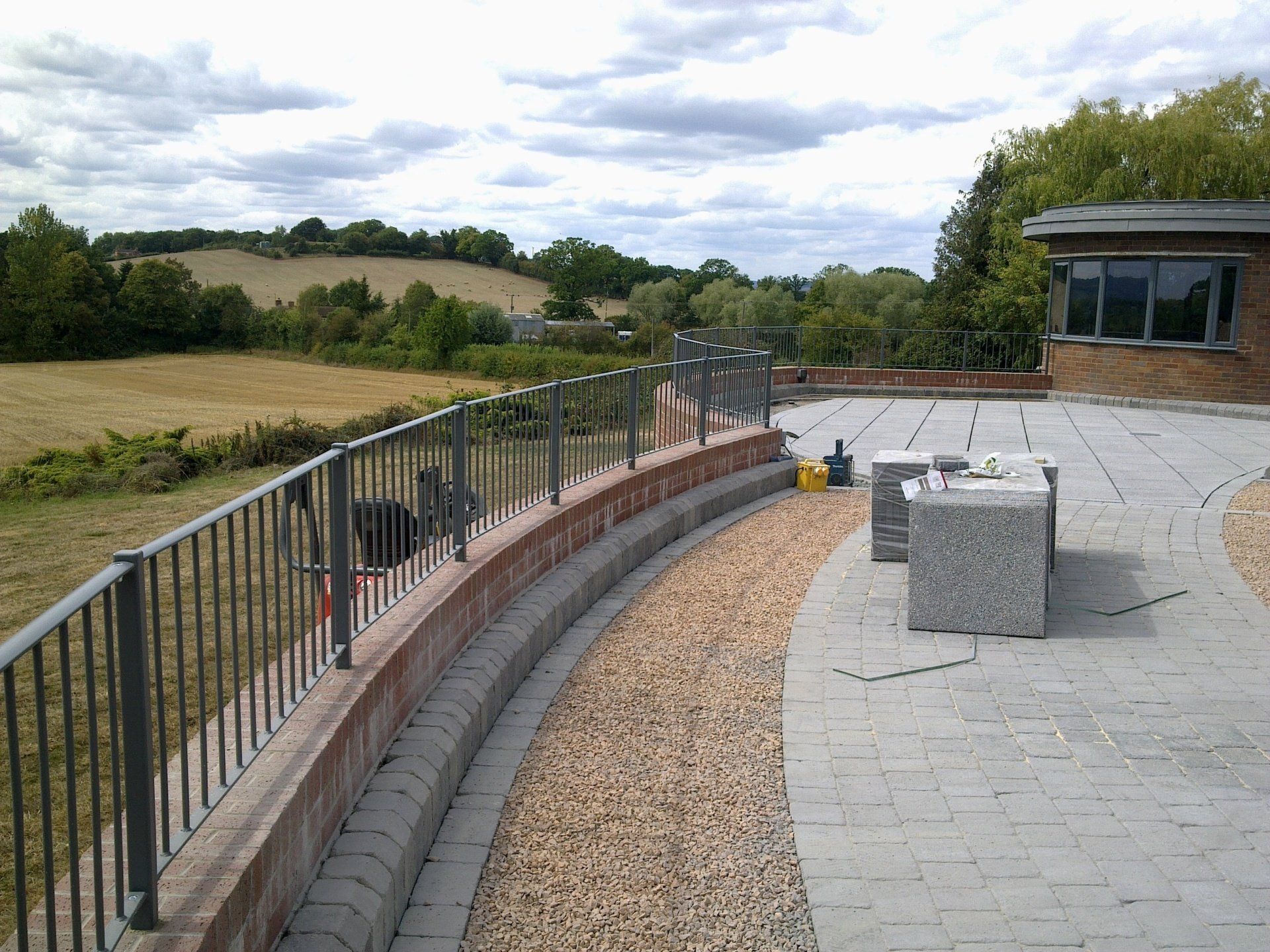 Curving brick wall with metal railing, paved patio, gravel border, and field in the background under a cloudy sky.