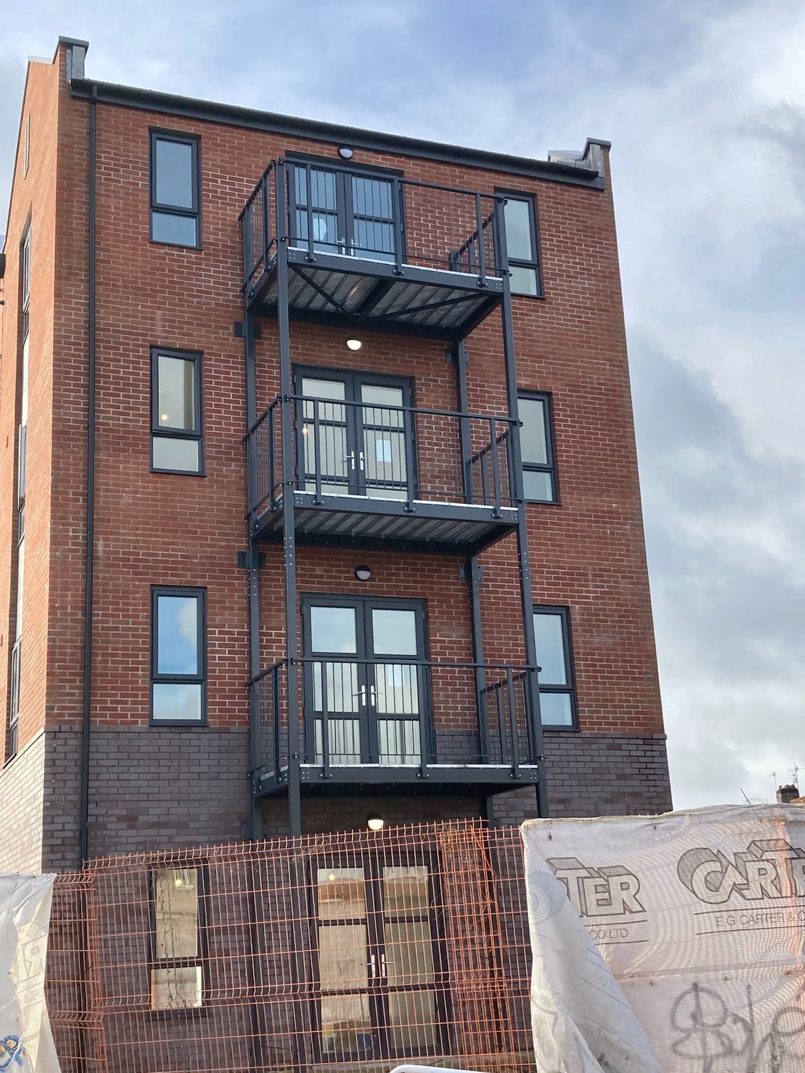 Red brick building with exterior metal balconies and black-framed windows; cloudy sky.