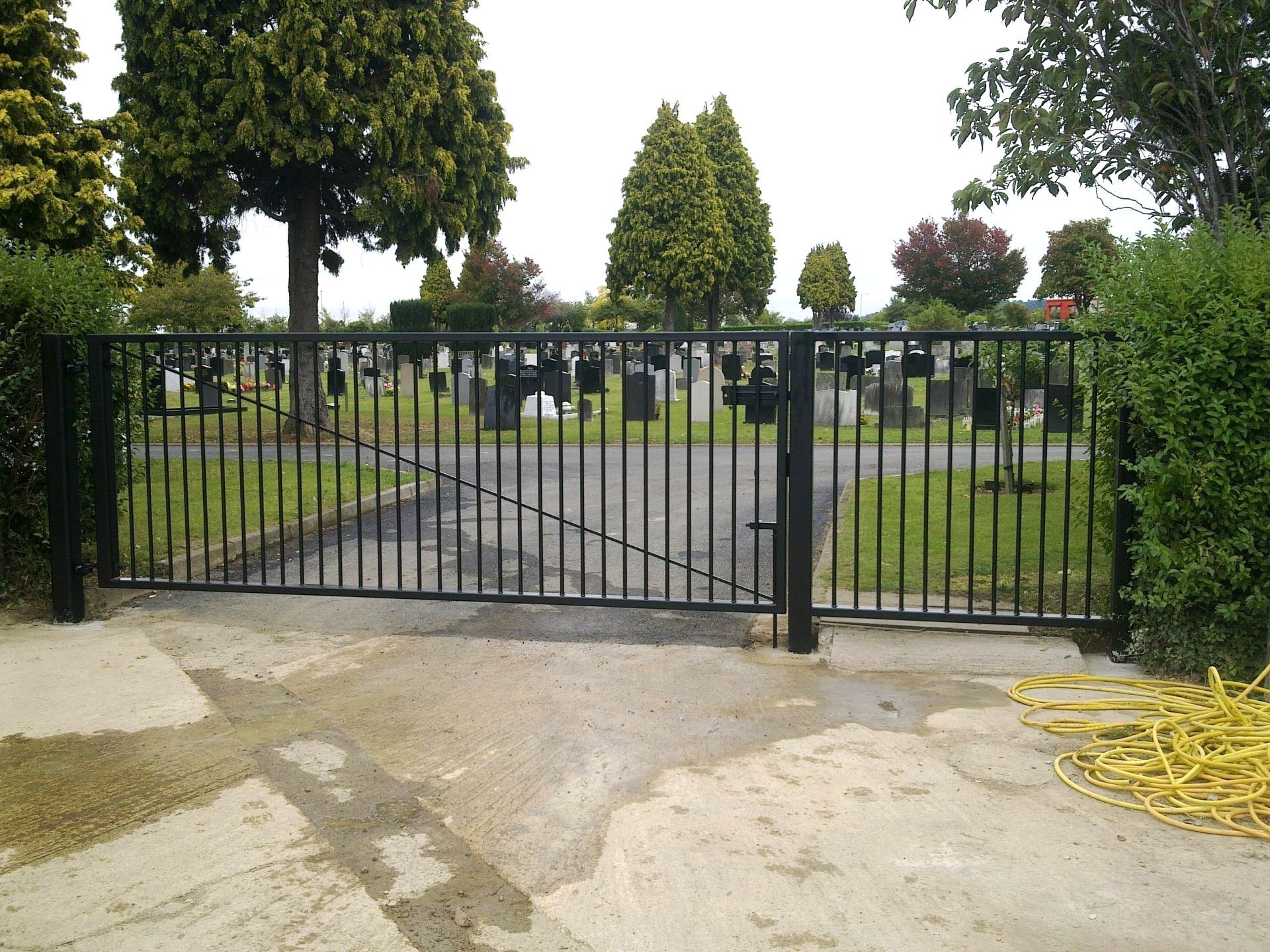 Black metal gate, open, at a cemetery entrance; gray tombstones, green grass, trees.