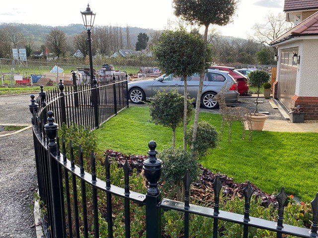Black iron fence borders a manicured lawn with topiary trees and parked cars.