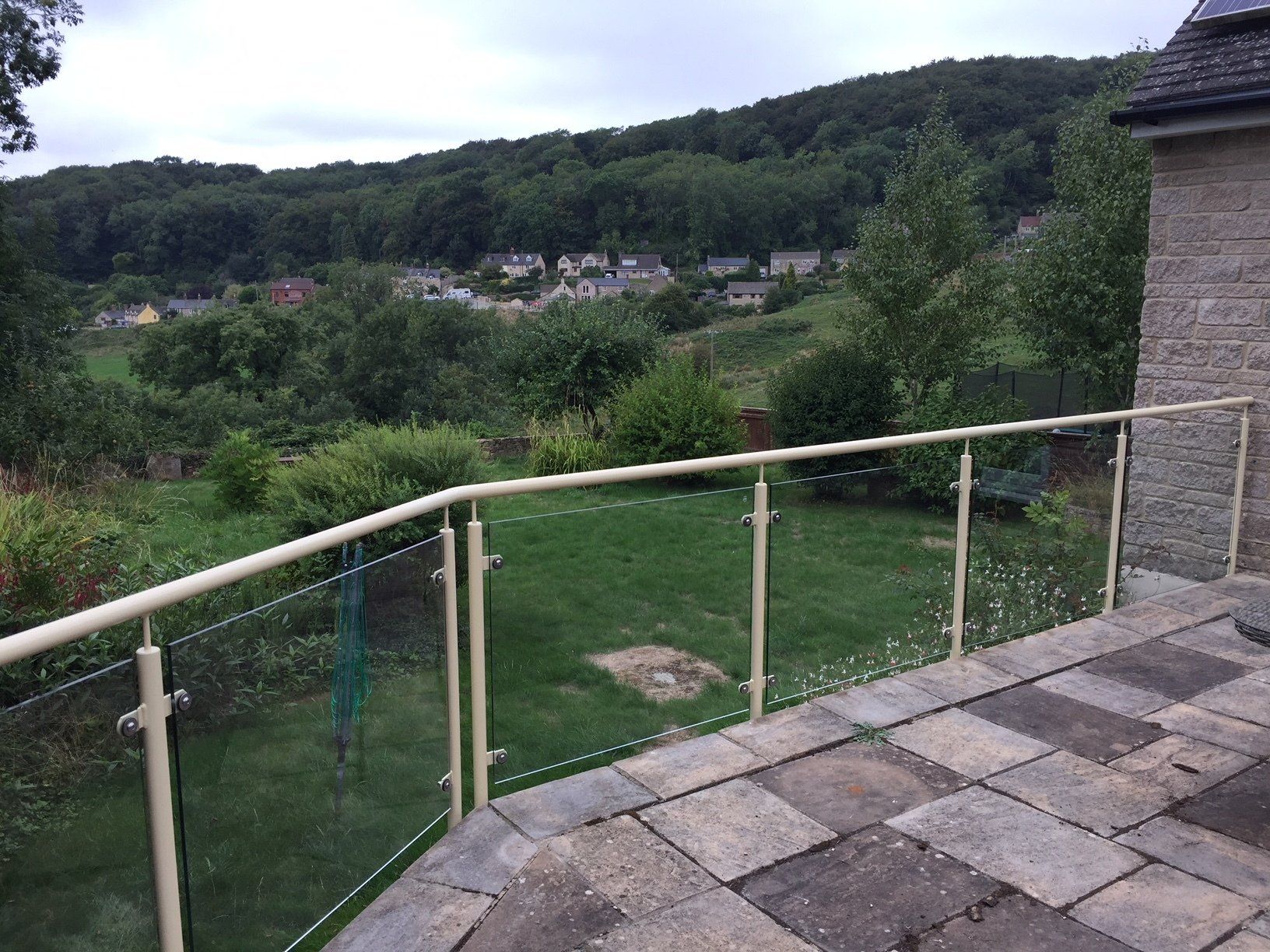 Patio with glass railing overlooking green yard and distant trees. Cloudy sky.