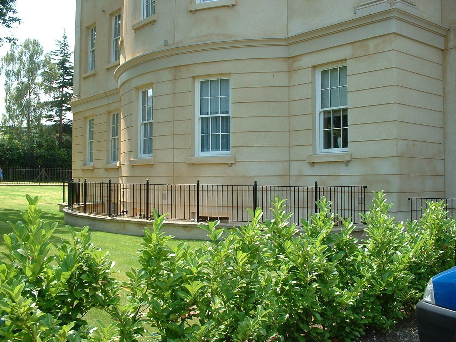 Curved, light-colored building with multiple windows. A low black fence separates the building from a hedge and lawn.