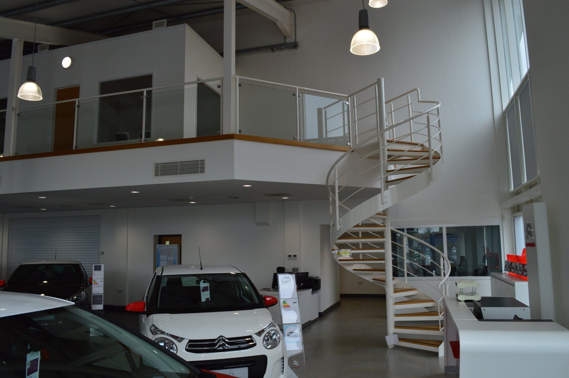Car dealership interior with cars on display, white walls, mezzanine level, and spiral staircase.