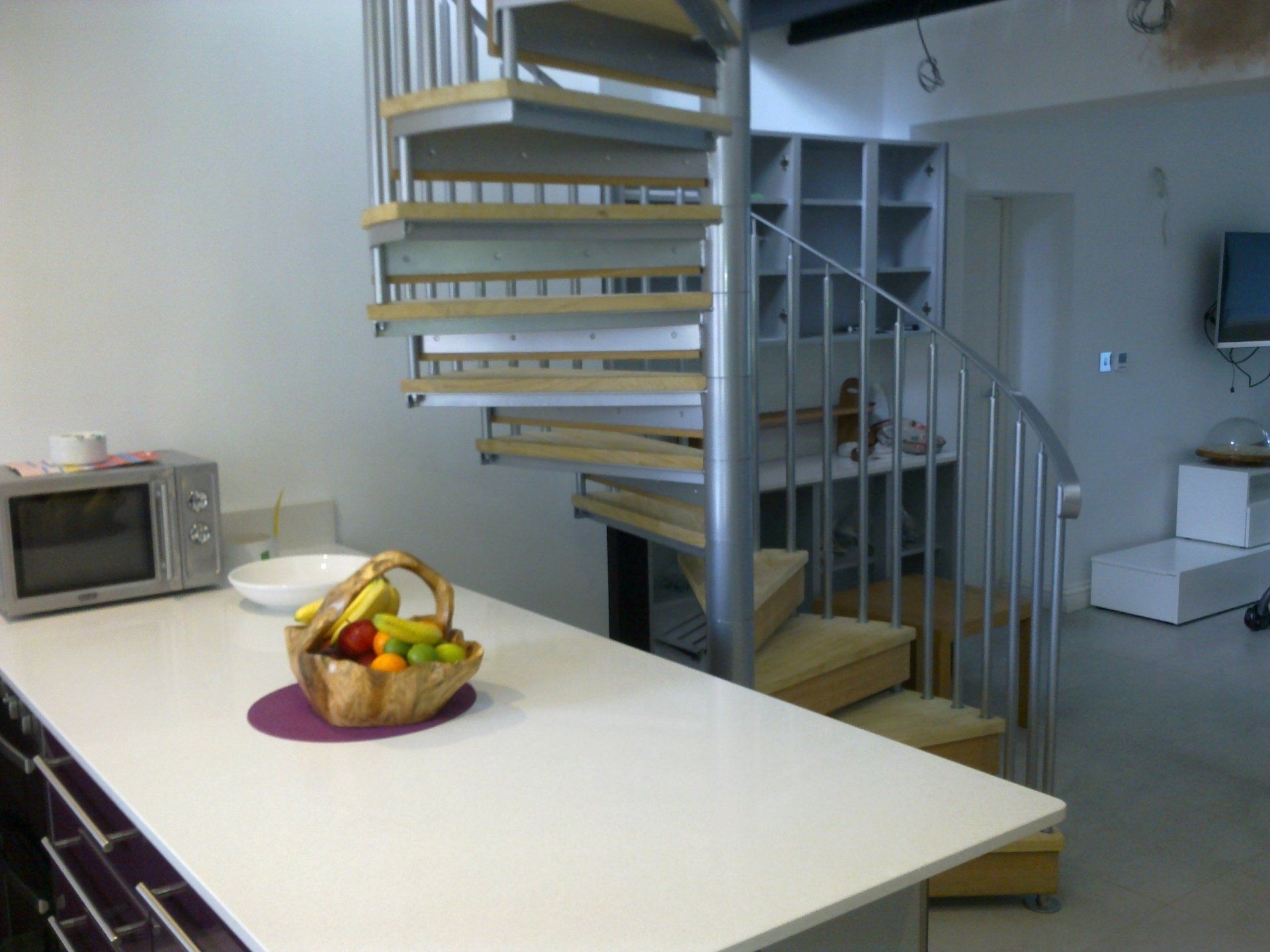Kitchen with white counter, fruit basket, microwave, and a spiral staircase.