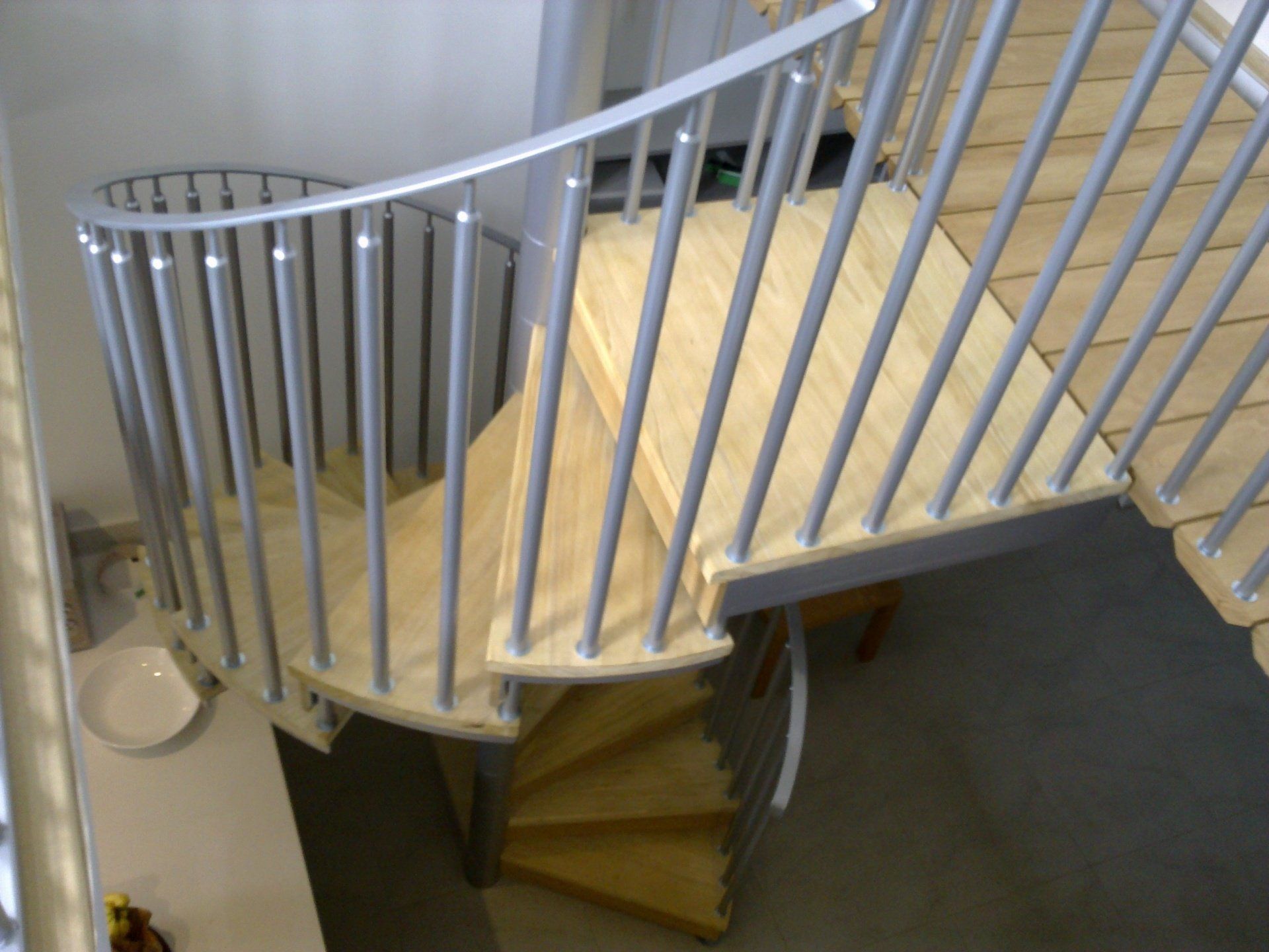Light wood spiral staircase with silver railings and vertical supports.