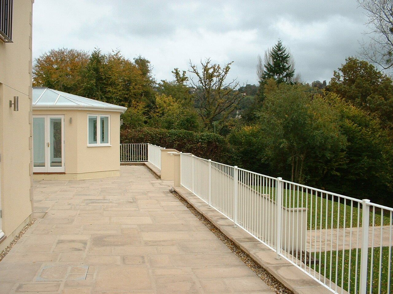 Patio with cream-colored building and white railing overlooking trees under a cloudy sky.