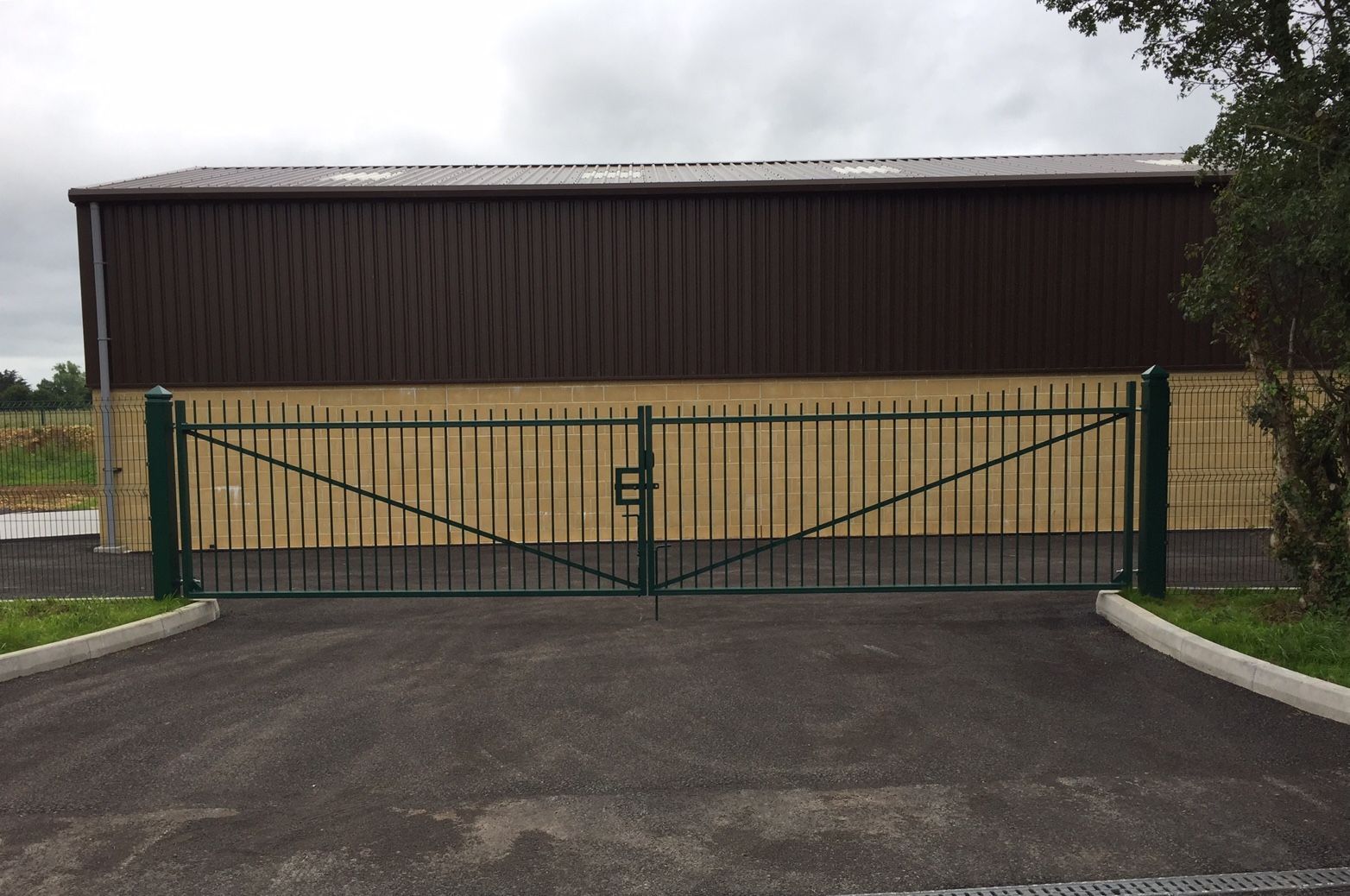 Green metal gate in front of a brown and tan building with an asphalt driveway.