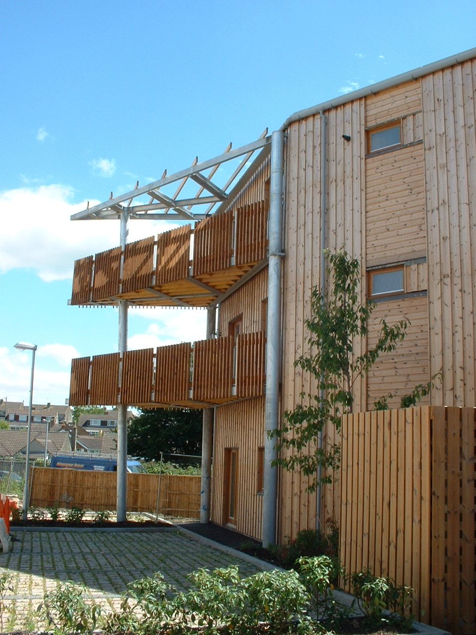 Wooden building with balconies and a glass roof, with a brick driveway and green bushes.