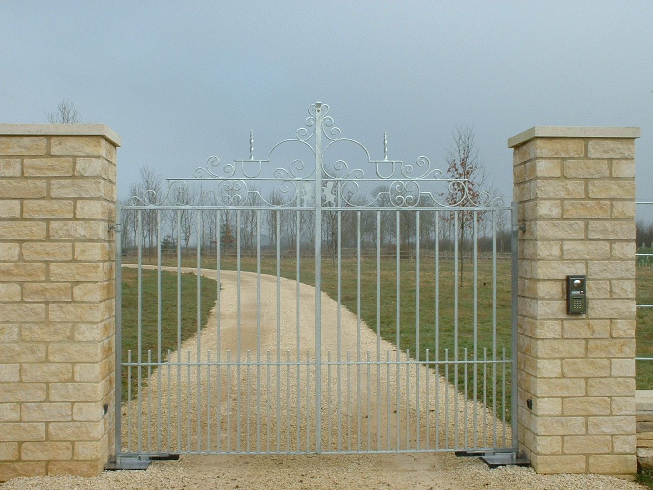 Metal gate between brick pillars, opening to a gravel driveway.