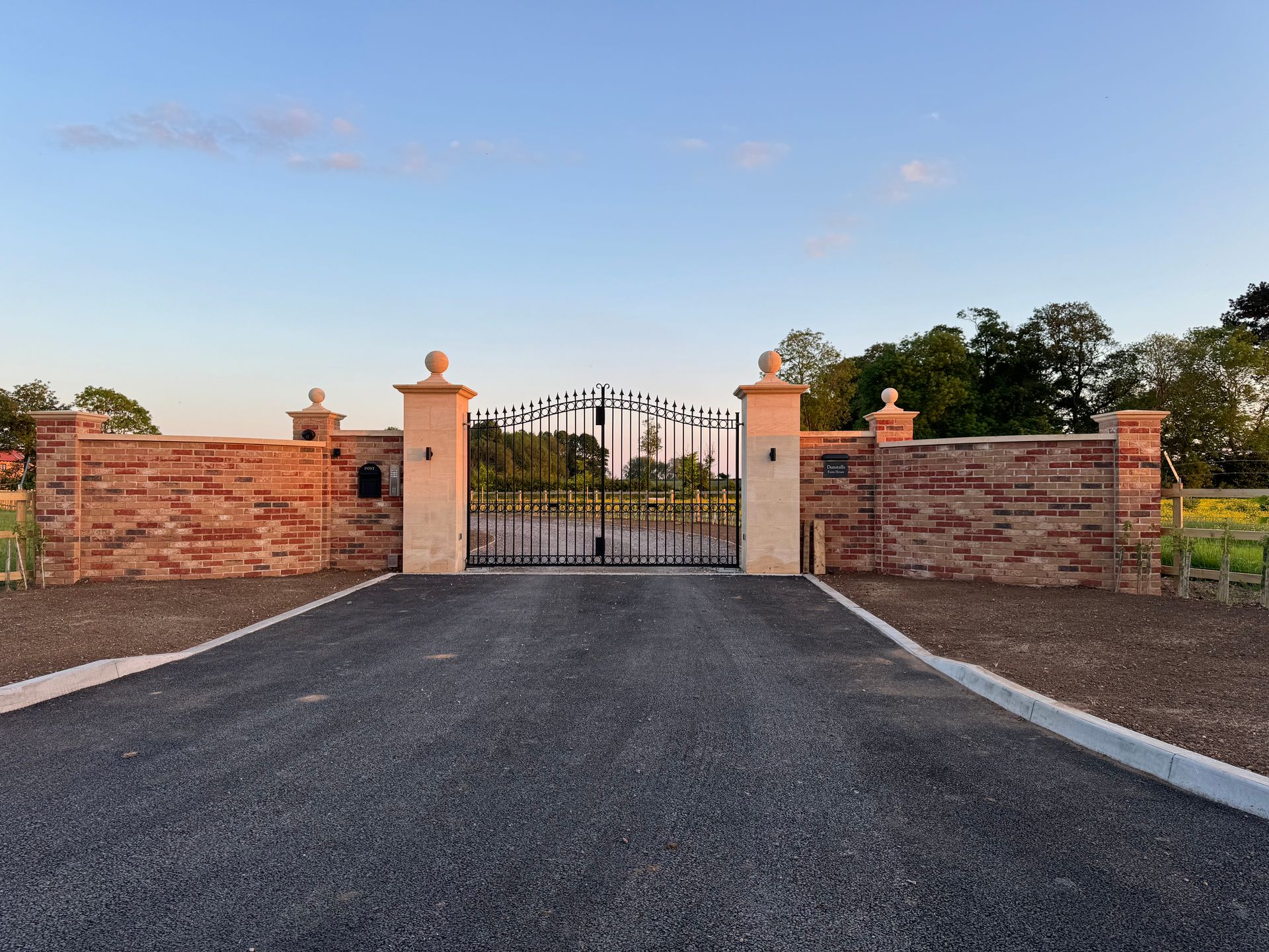 Red brick wall and ornate black gate on a paved driveway, topped with decorative pillars against a blue sky.