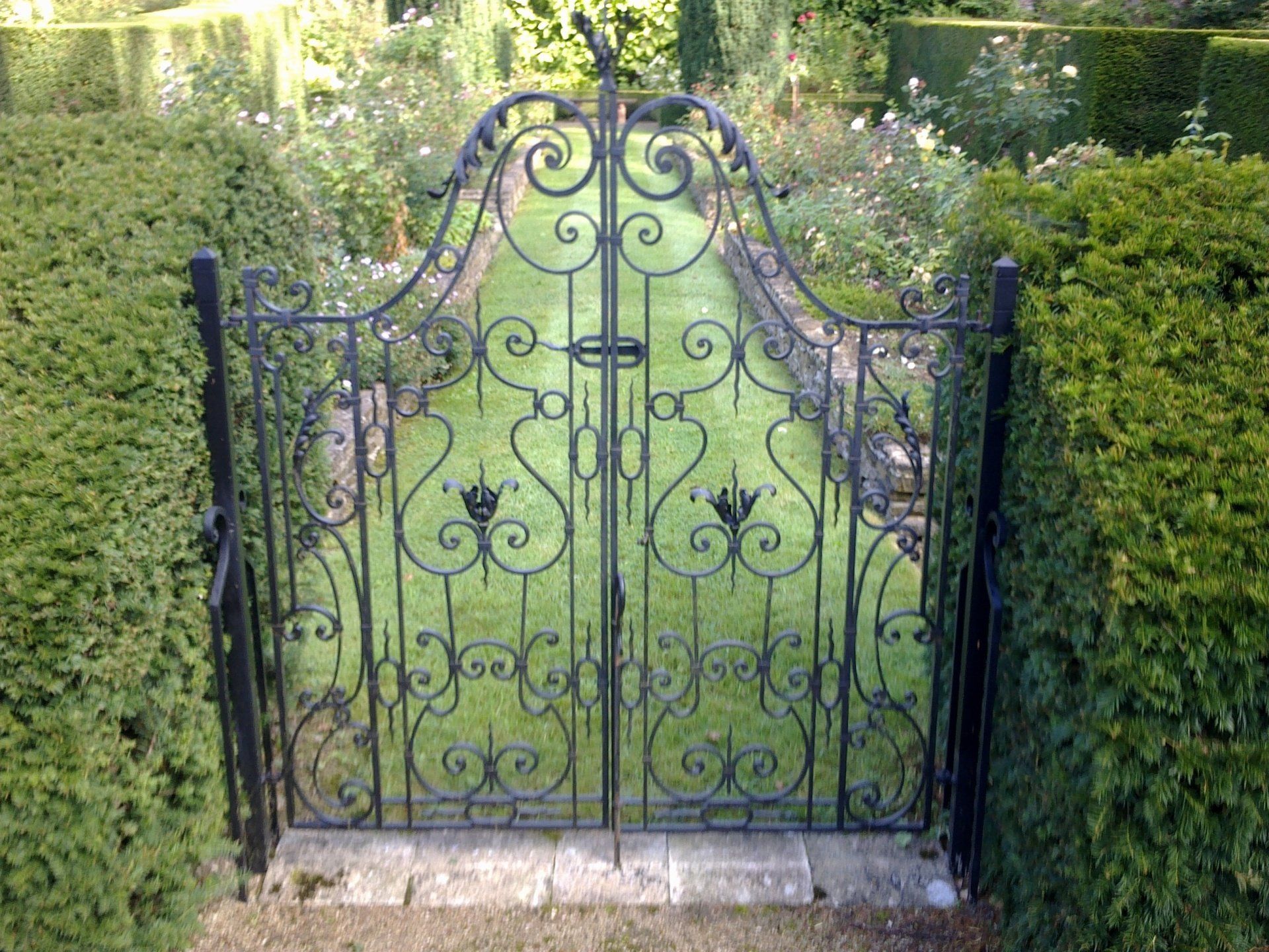 Black ornate wrought iron garden gate set in trimmed hedges, leading to a path.