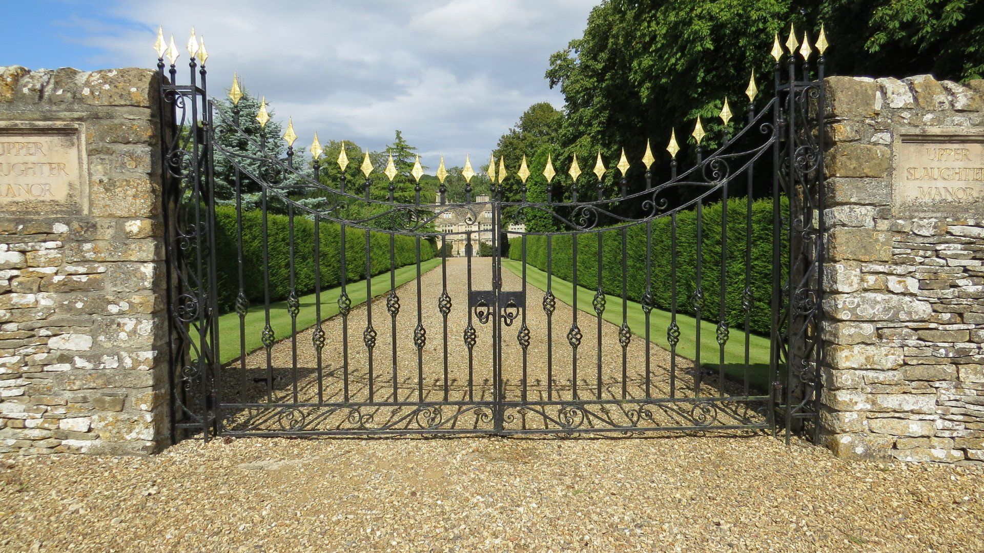 Black wrought-iron gate with gold finials, set between stone walls, opening onto a gravel path lined with hedges.