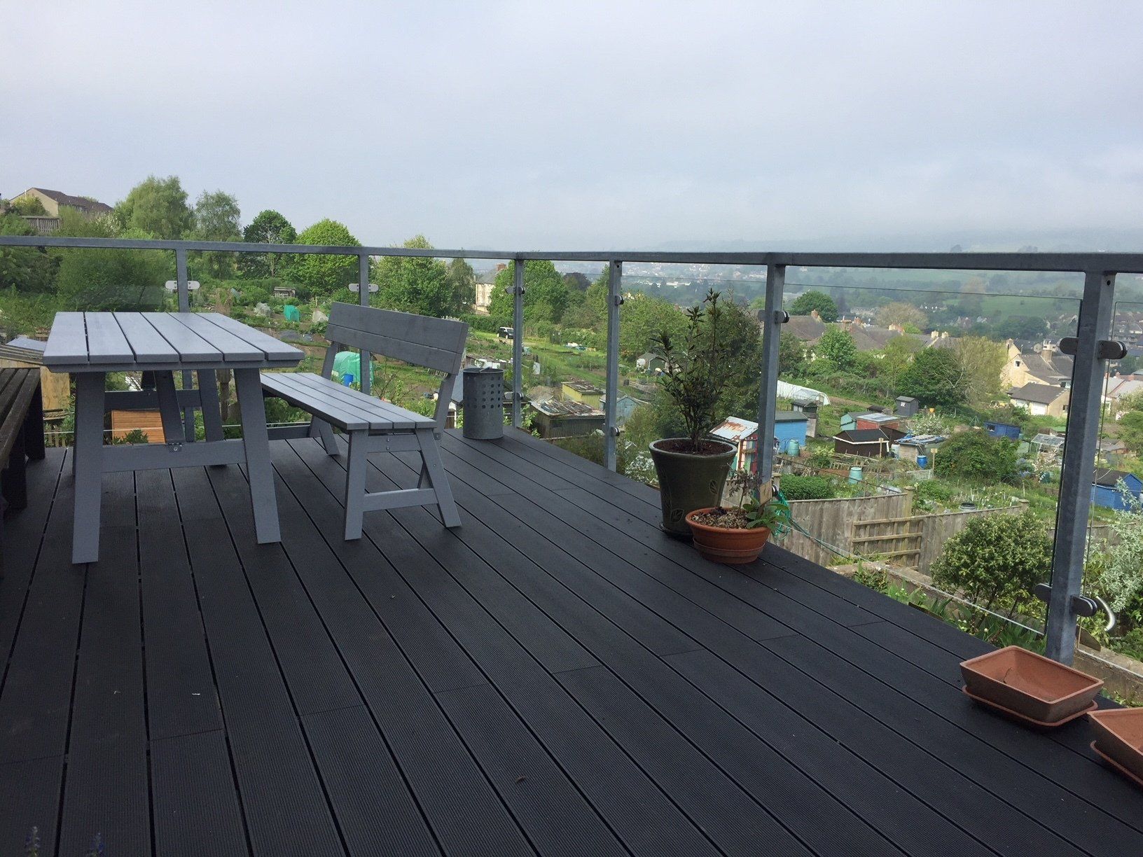 Gray outdoor deck with table, bench, and plants overlooking a town.