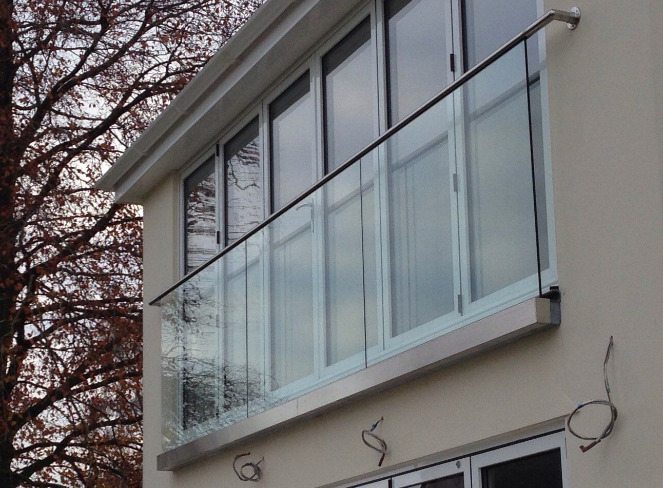 Balcony with glass panels and a silver railing on a beige building with a leafless tree in the background.