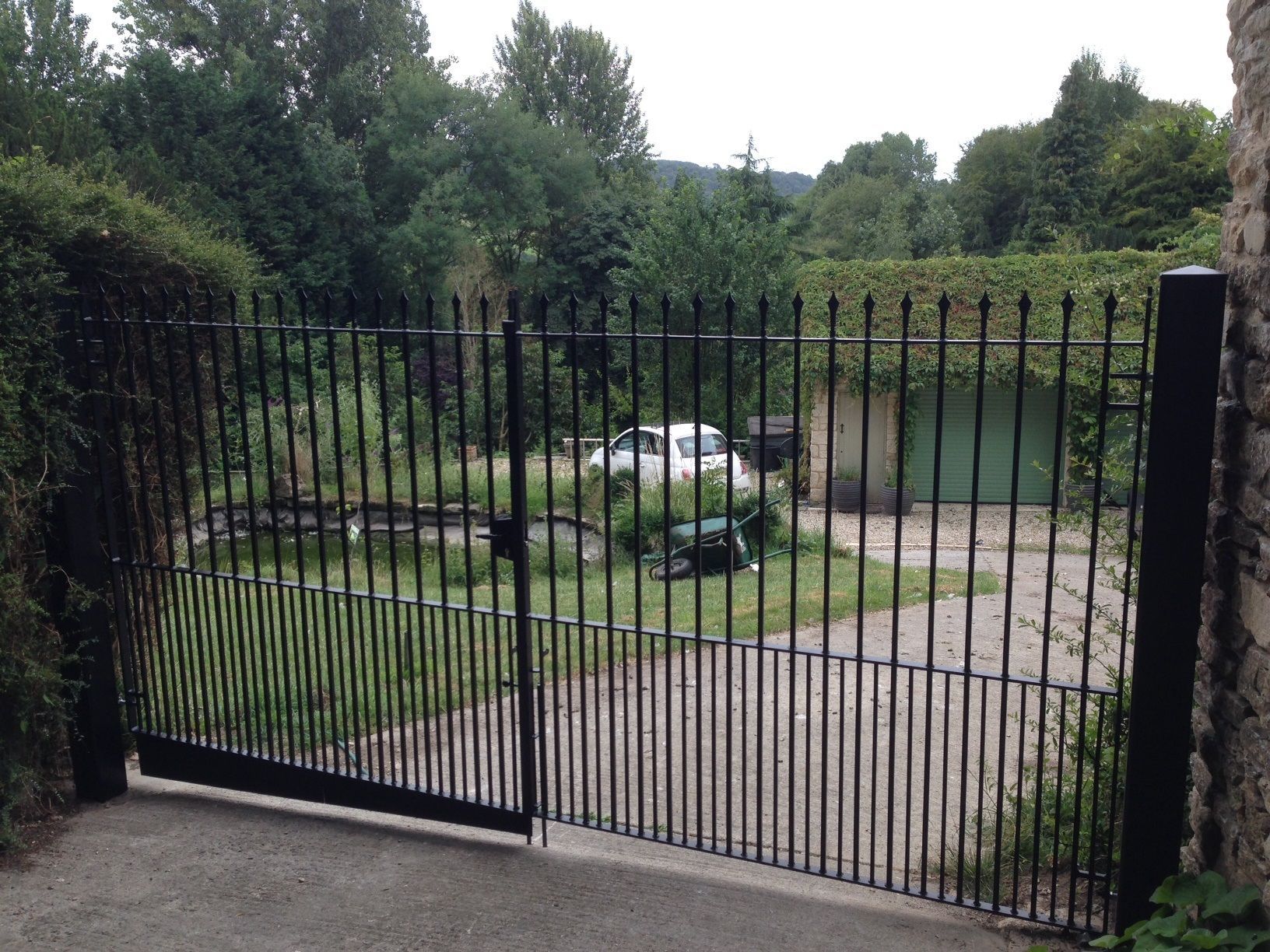 Black metal gate open, leading to a driveway with a white car and overgrown greenery.