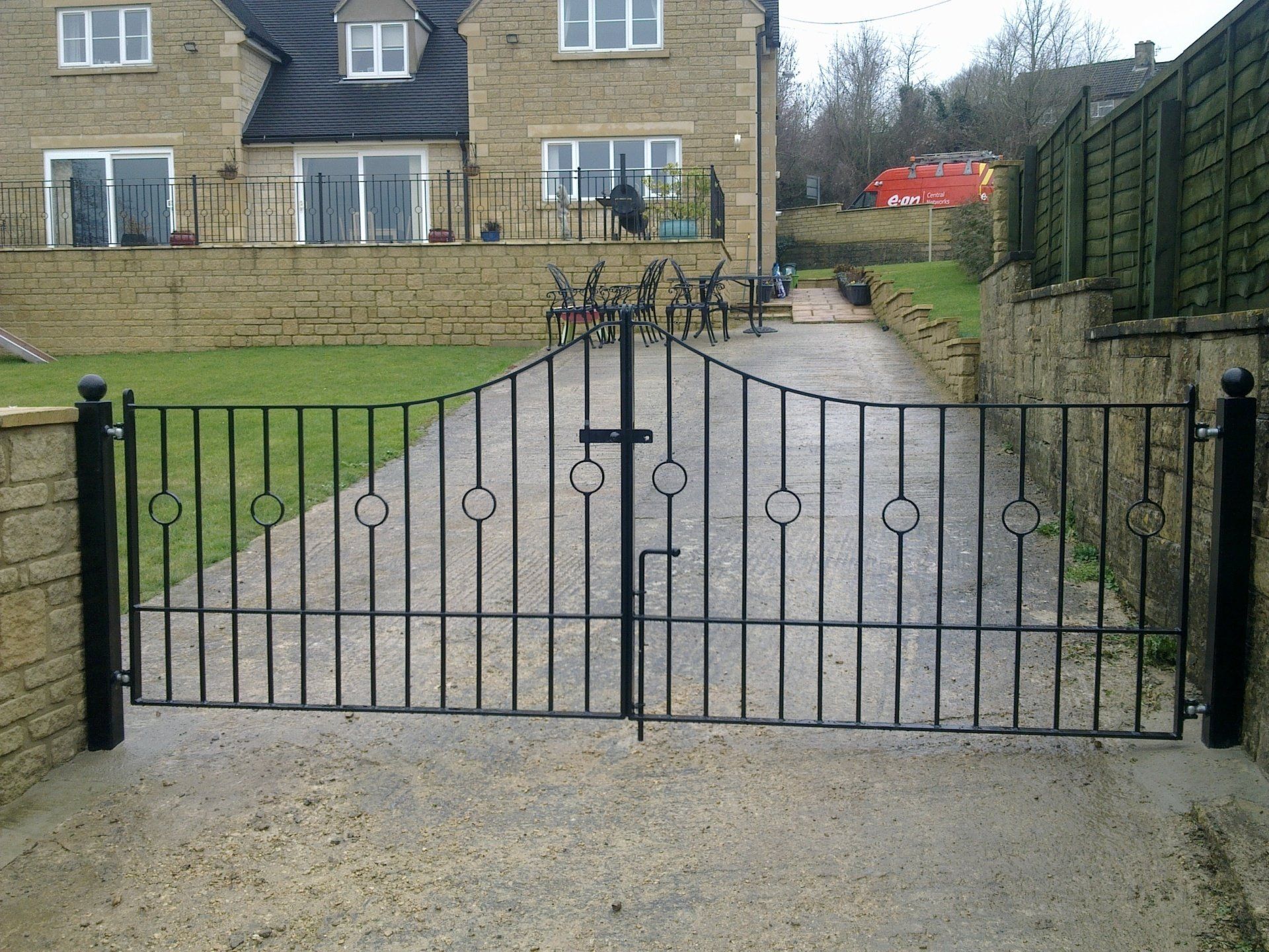 Black metal driveway gates in front of a light-colored stone house and a concrete driveway.