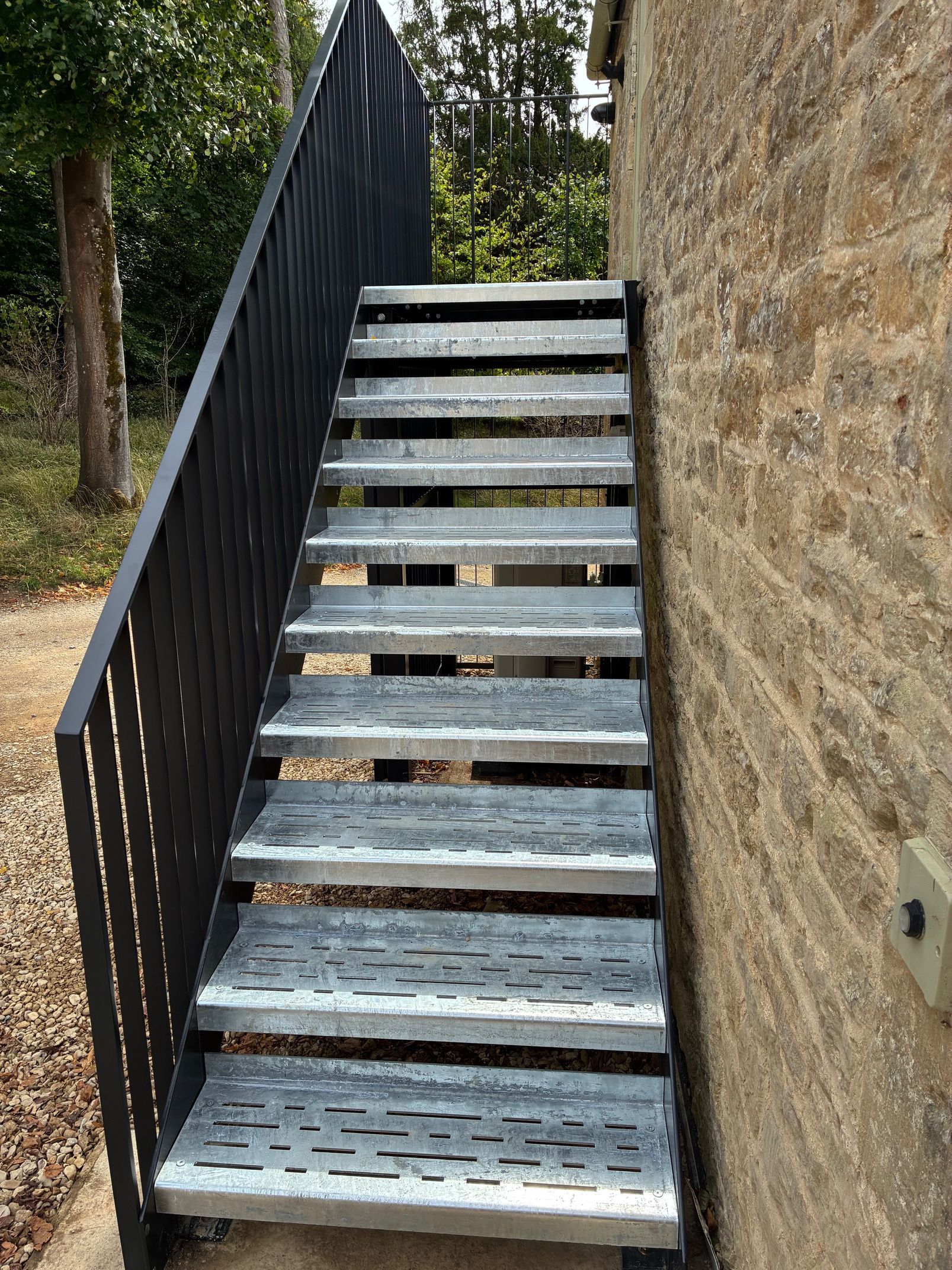 Metal staircase with black railing next to a stone wall; outside.