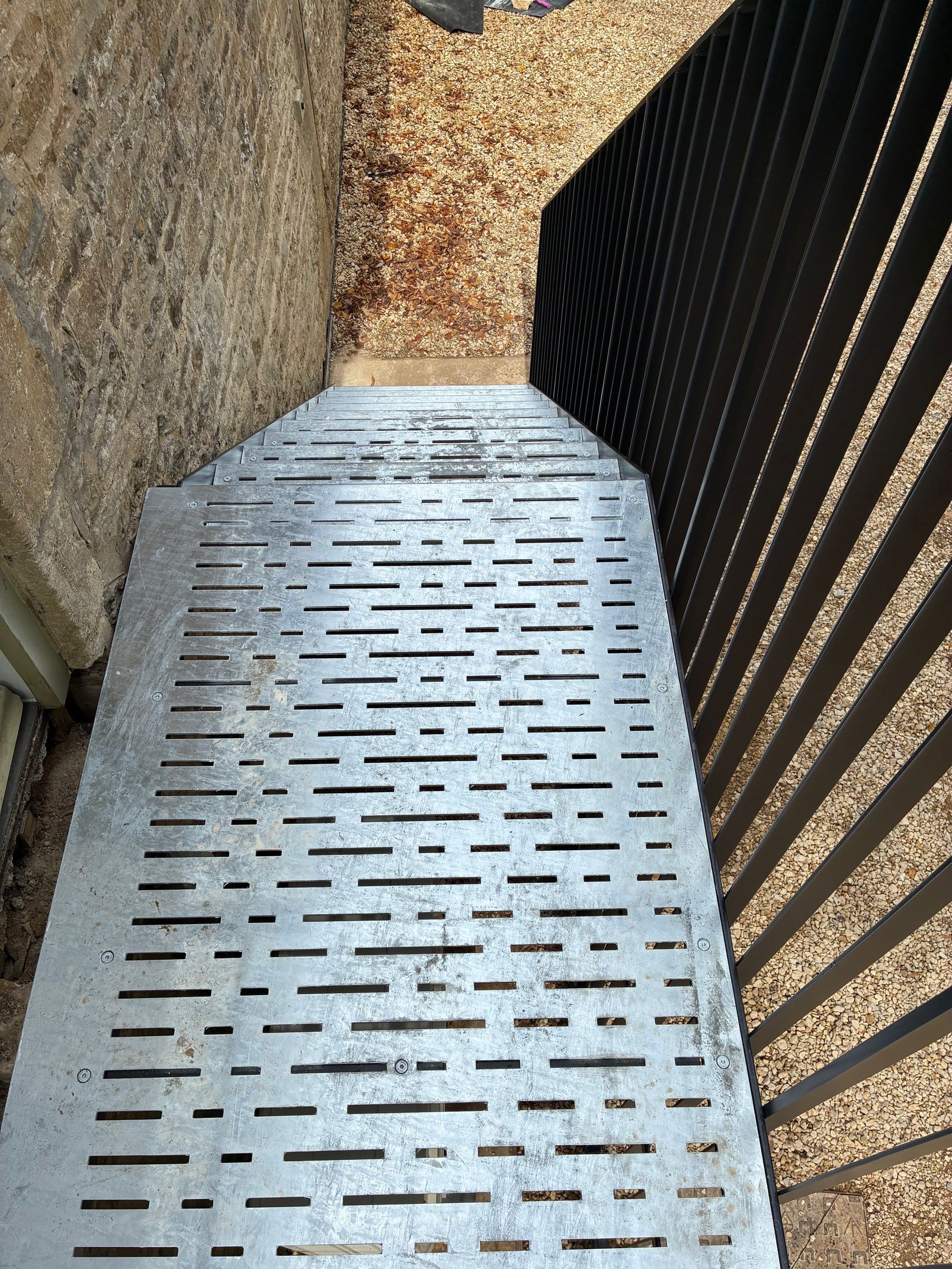 Metal ramp with slotted pattern leads to a doorway; brown wall and black metal fence frame it.