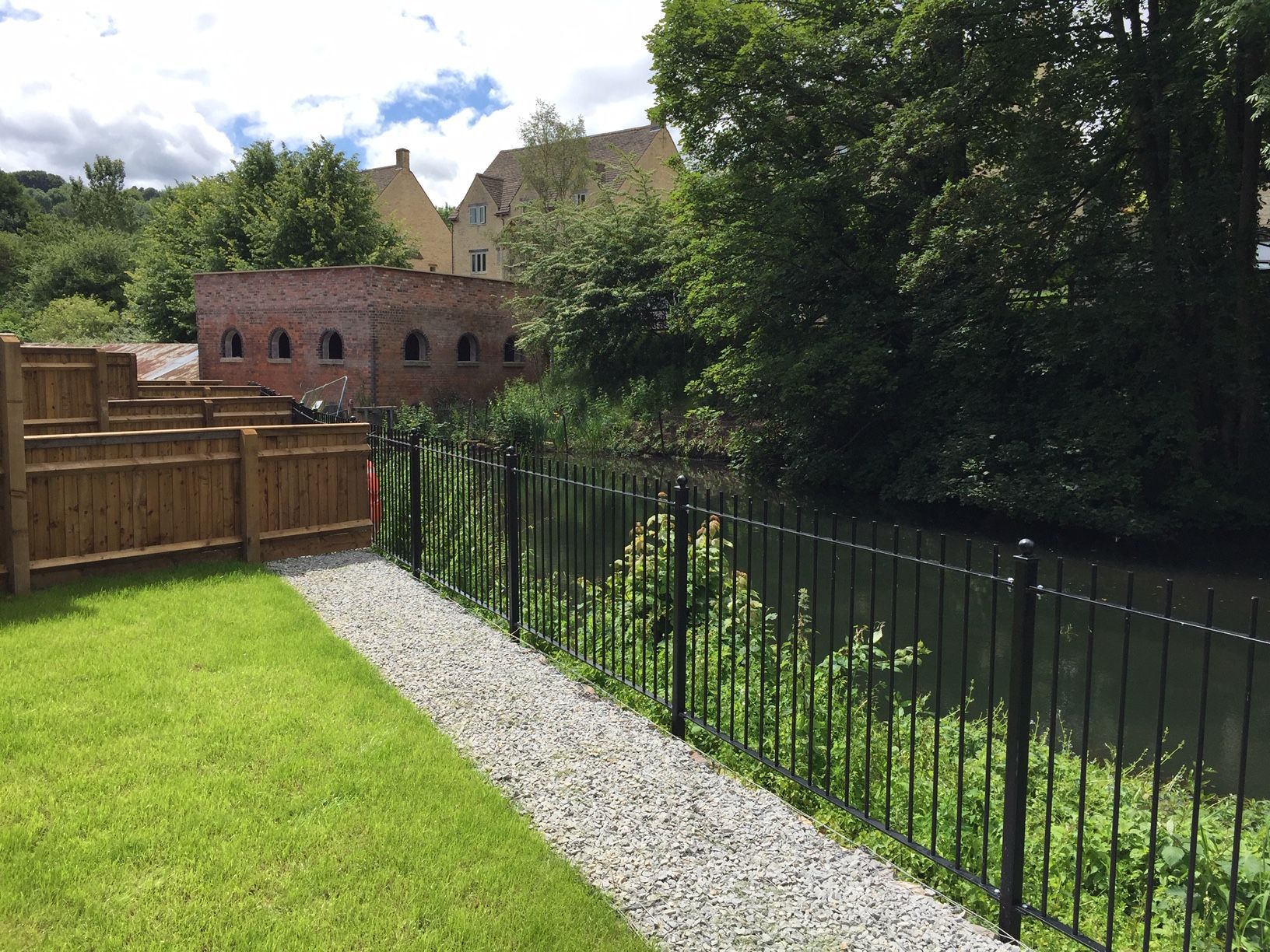 A grassy yard with a gravel path and black fence borders a river, with a brick building in the background.
