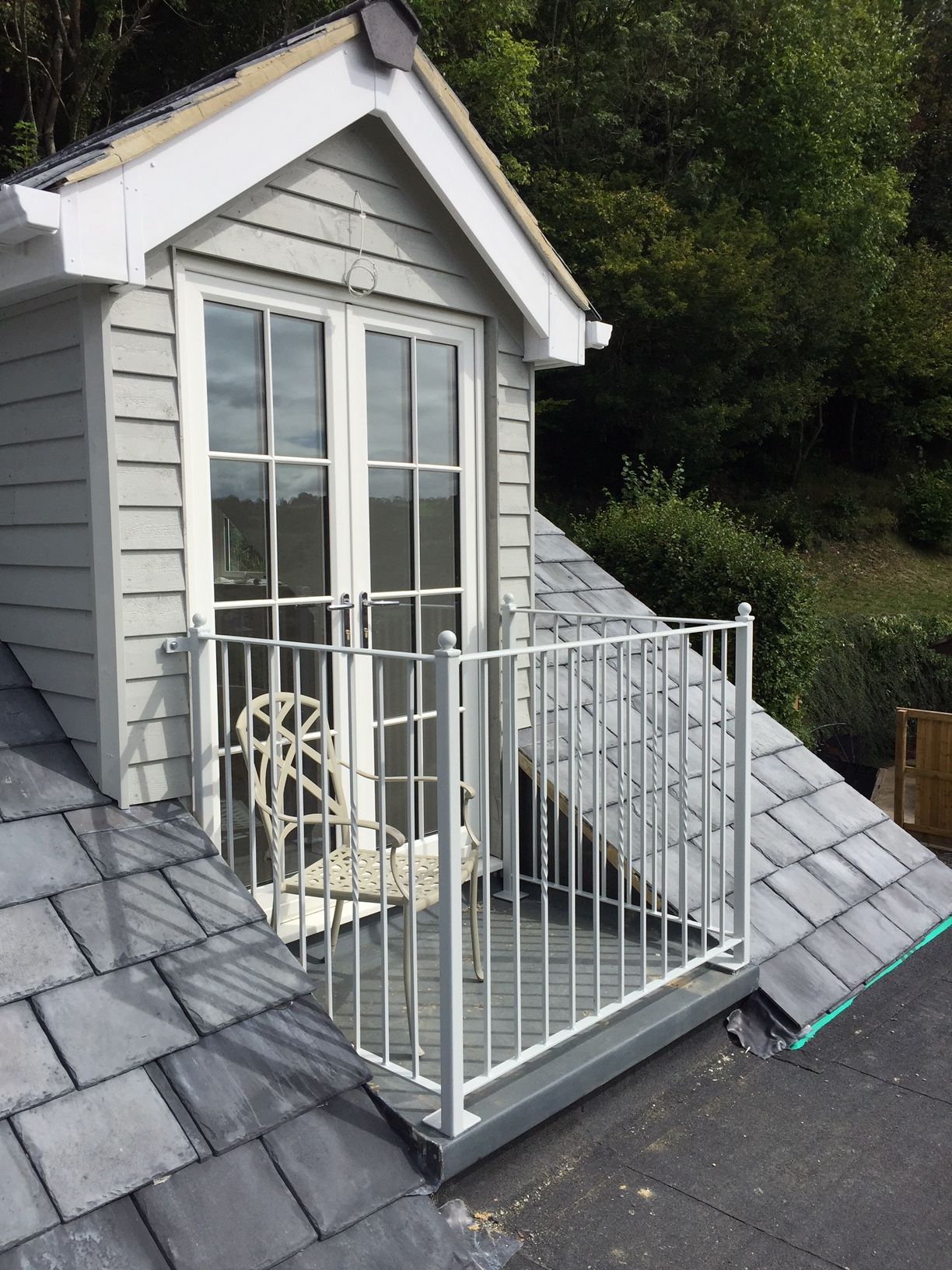 Dormer with a small balcony, white railing, gray siding, and dark roof tiles; set against trees.