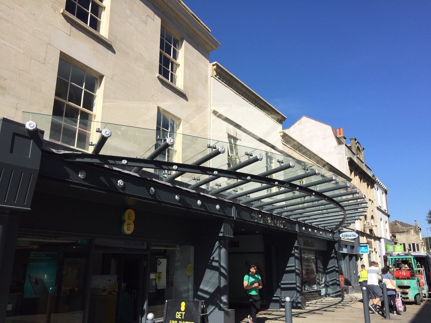 Storefront with glass awning on a sunny street. People walking nearby; bright sky.