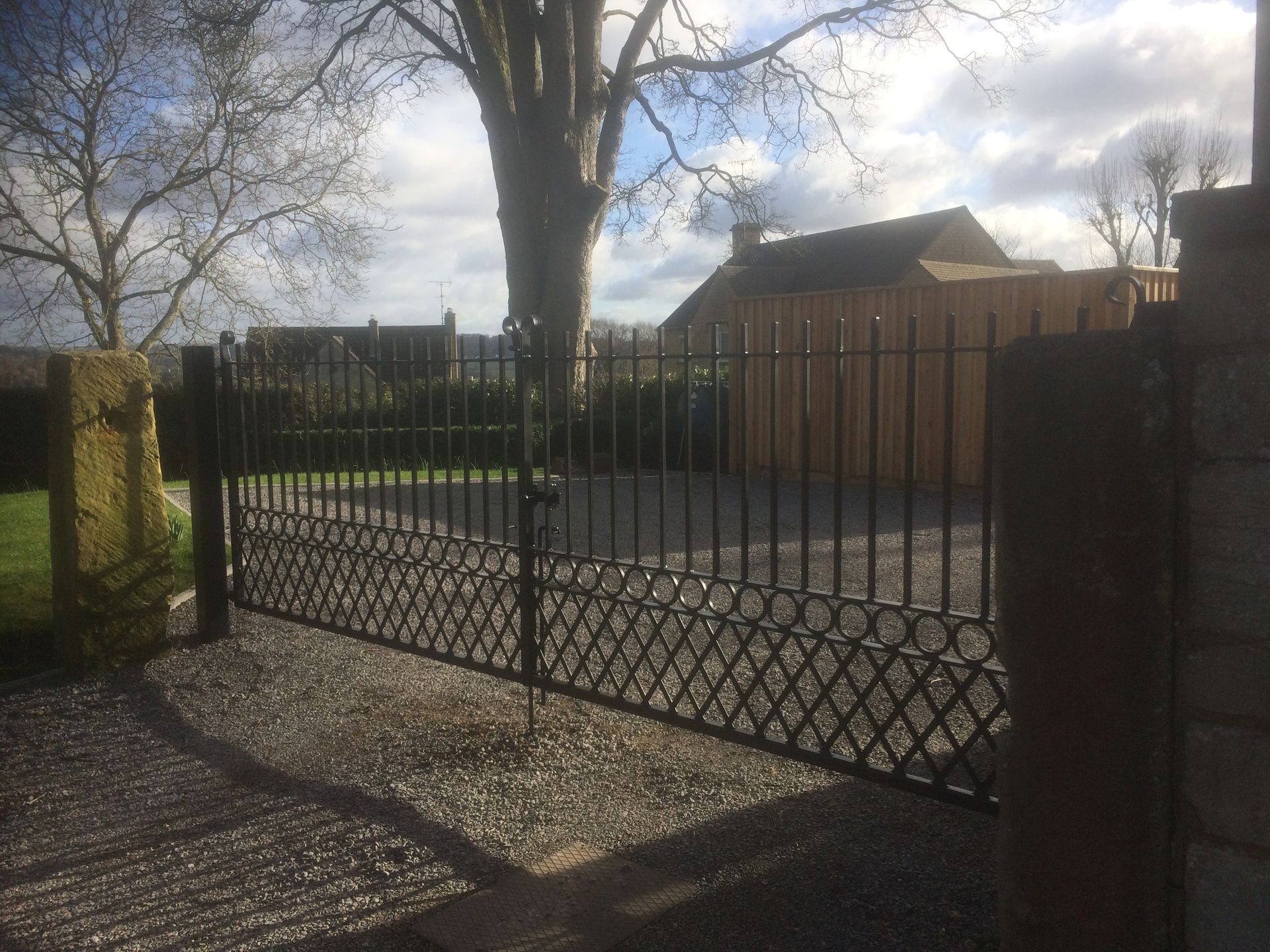 Black metal gate, gravel driveway, leading to a wooden shed and tree. Sunny day.