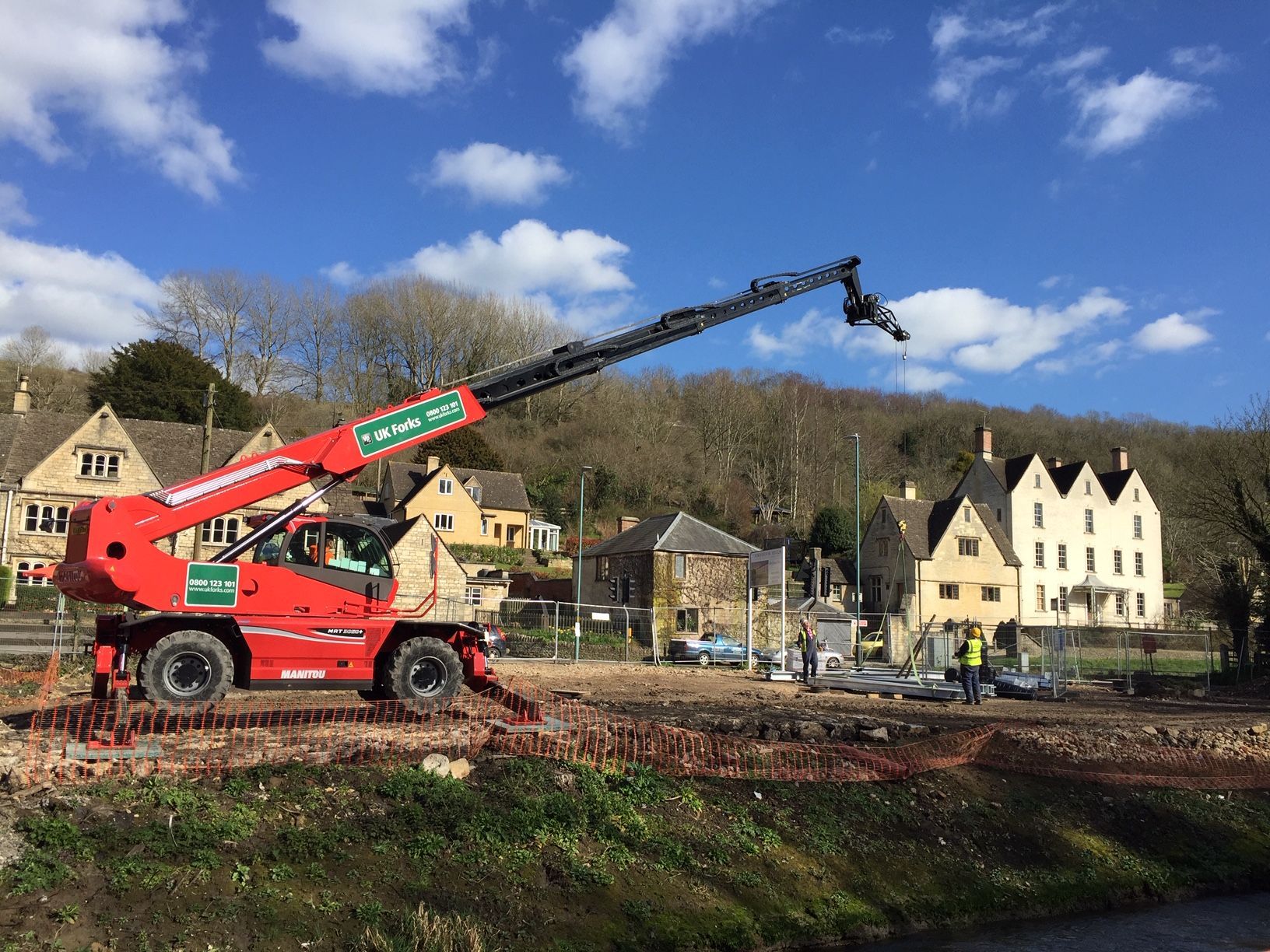 Red telehandler on a construction site with buildings and trees in the background under a blue sky.