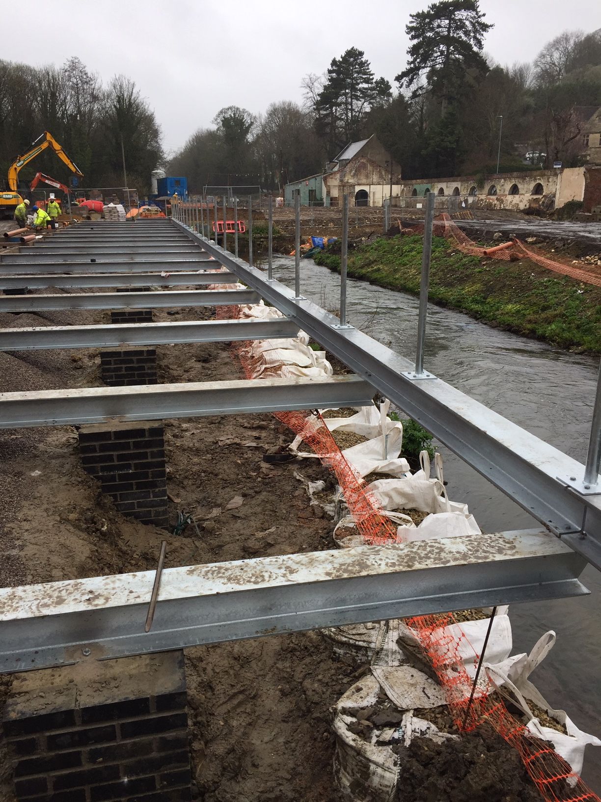 Construction site with metal beams over a waterway. Workers and an excavator are visible in the background.