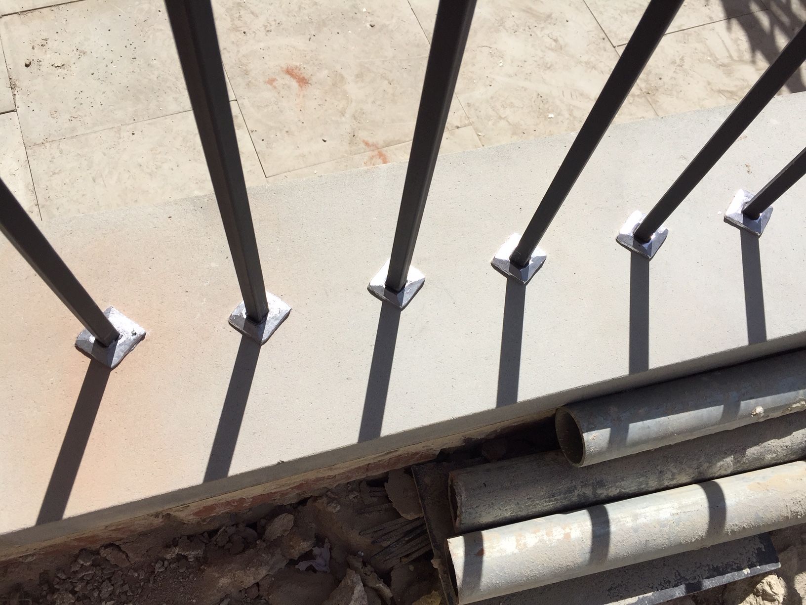 Metal railing posts anchored to a concrete surface, casting long shadows. Construction materials nearby.