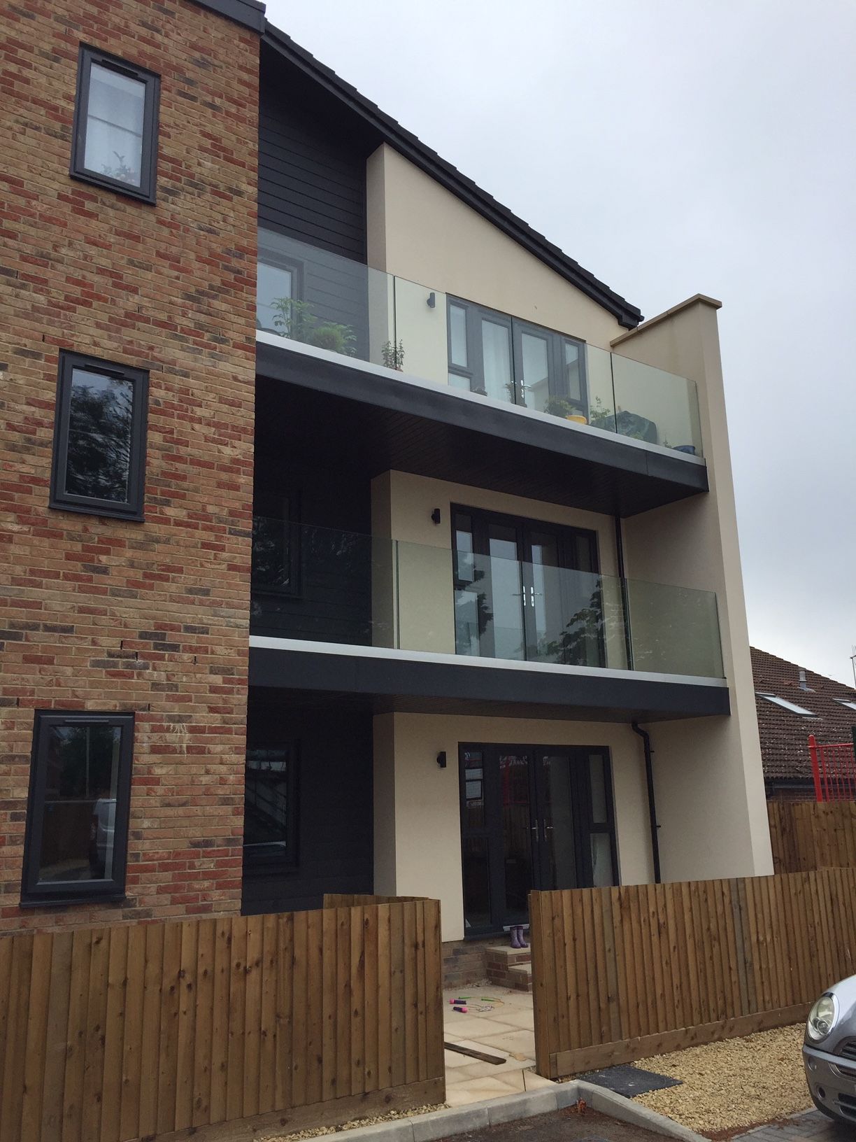 Modern apartment building with brick, dark siding, and glass balconies. Brown wooden fence in front.