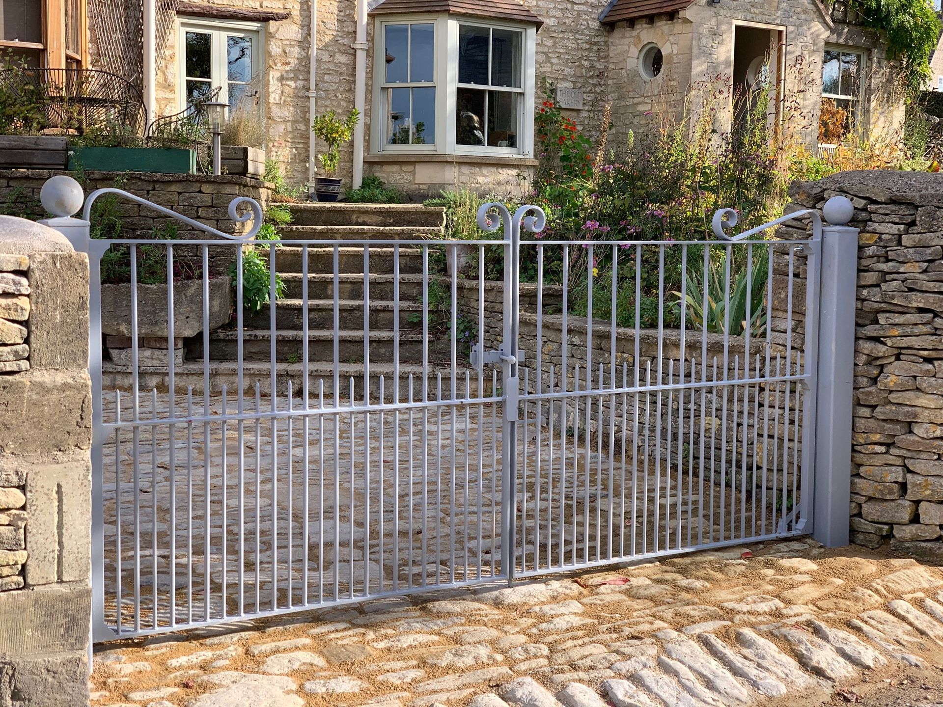 Metal gate in front of a stone house; a double gate with vertical bars, gray in color, opening onto a path.