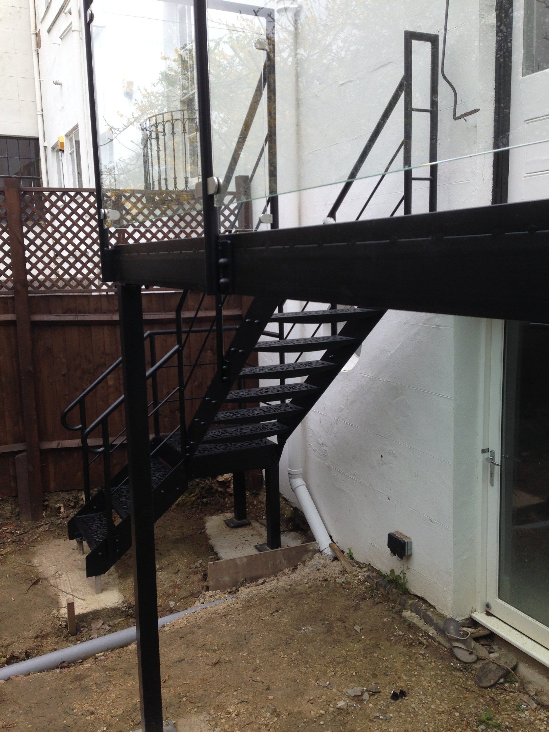 Black metal exterior staircase leading up to a platform. Stairs set against a white wall and dirt yard.